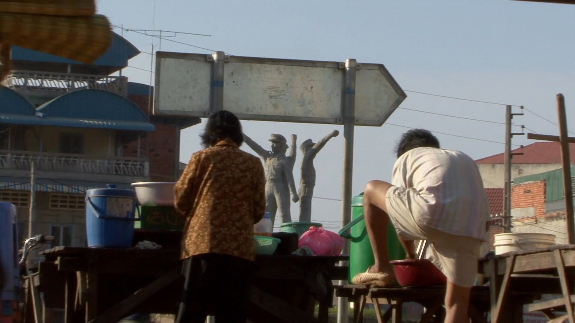 Daily scenes in a Cambodian town capture residents navigating public spaces near a prominent monument, culminating in a intimate moment of a mother nursing her child.