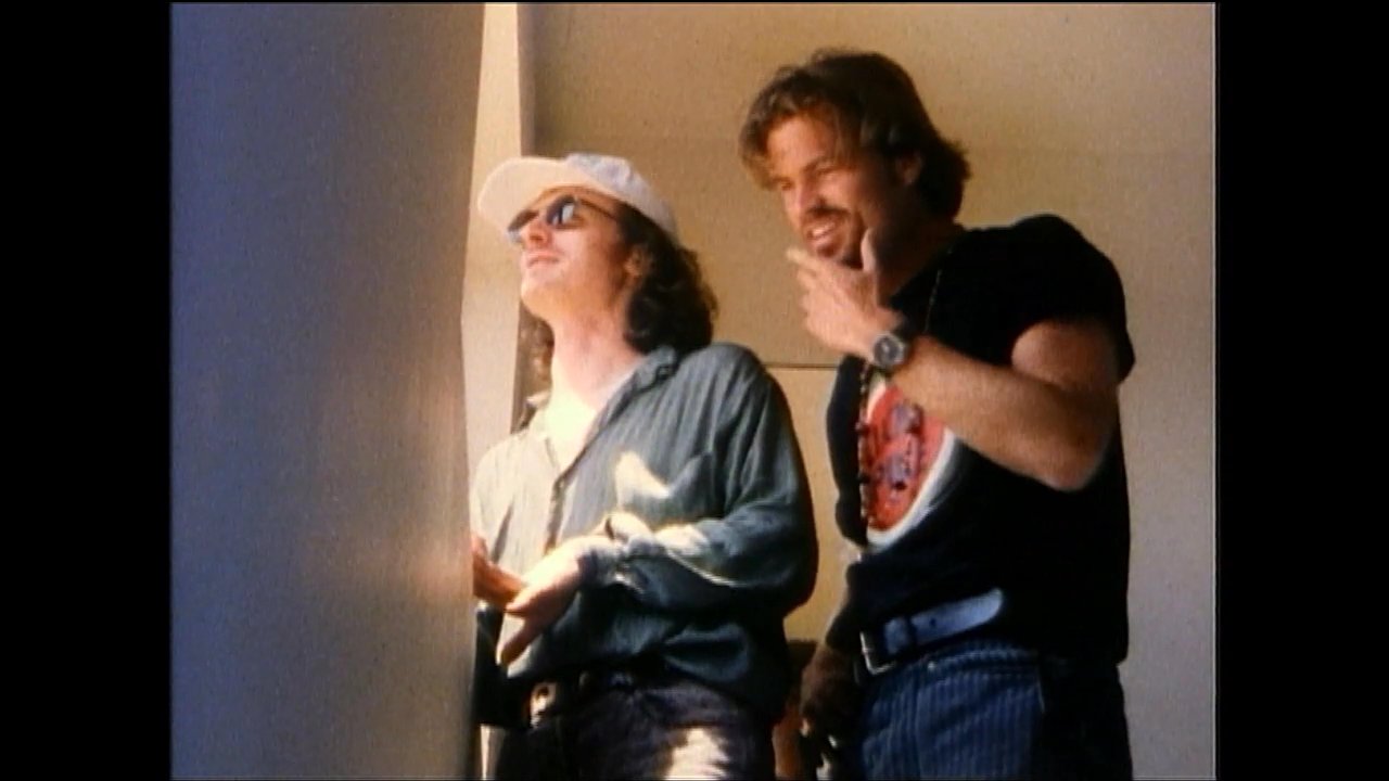 Two men engage in an animated conversation while standing against a wall in an indoor setting. One man gestures with his hands as he speaks to the other.