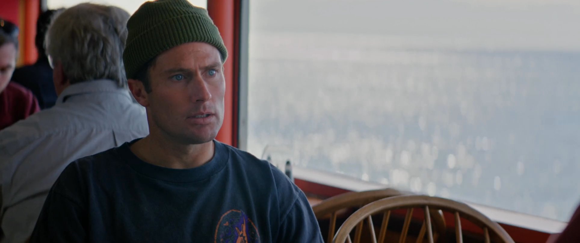 Two individuals engage in a serious, focused conversation while sitting on a ferry. The backdrop features sunlight reflecting off the water as they continue their discussion.