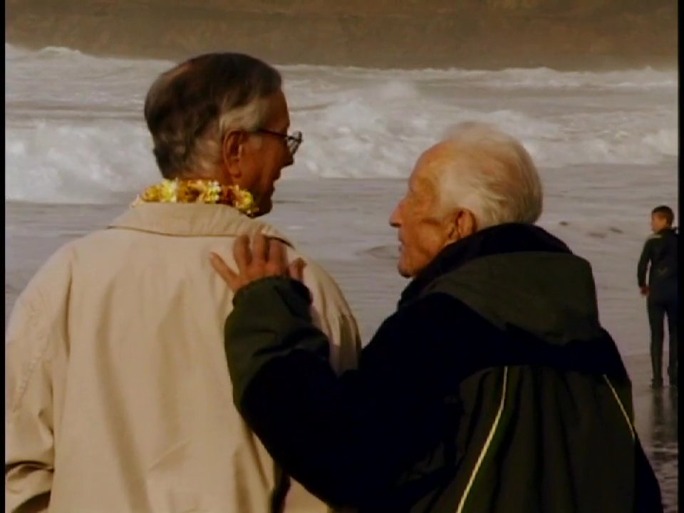 A group of senior friends shares stories and laughter over a meal before taking a group walk along the shoreline.
