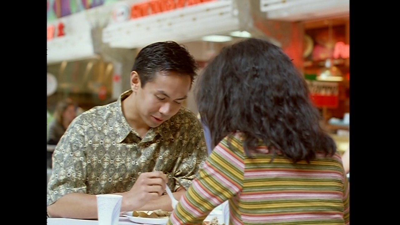 A couple engages in a focused conversation while sharing a meal at a bustling shopping mall food court. The scene transitions from the mall's common area to an intimate close-up of their table interaction.