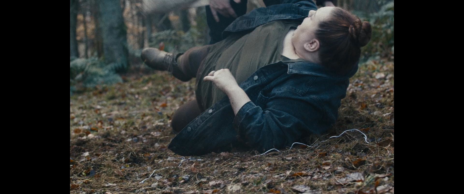 Two individuals engage in a violent physical struggle within a dense forest setting. The confrontation intensifies as they grapple on the forest floor, involving a canvas bag as a focal point.