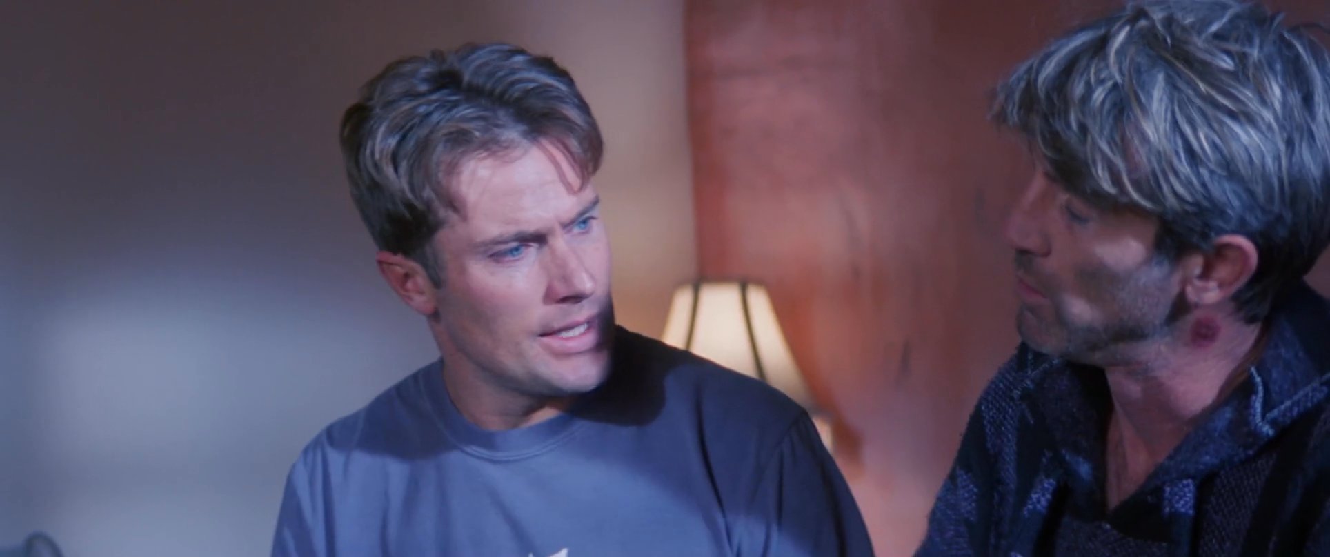 Two men engage in a serious, quiet conversation while sitting indoors, displaying visible tension and focus during their exchange.