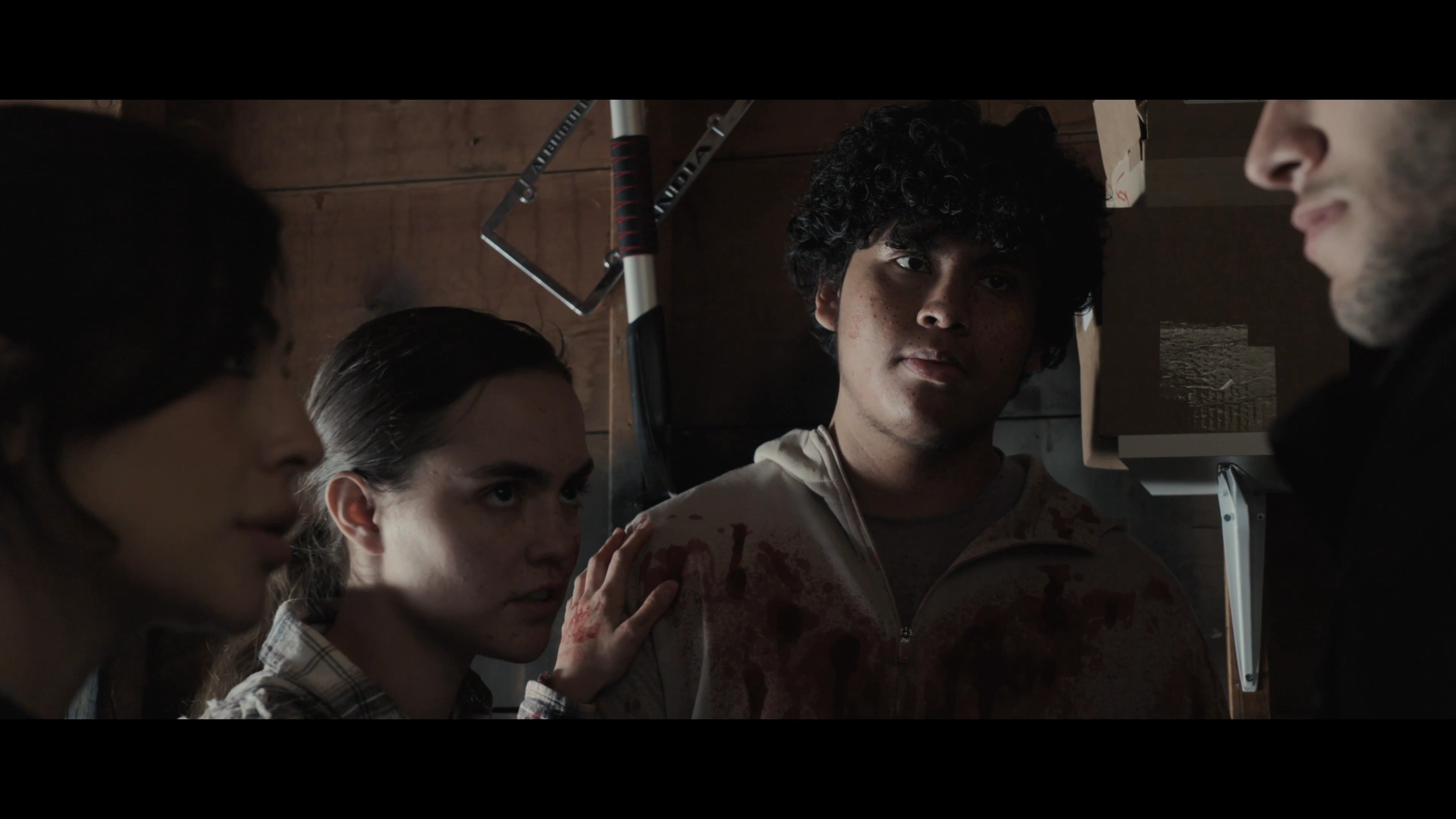 A group of young adults engages in a heated and tense conversation within a dark, wooden-walled garage setting.