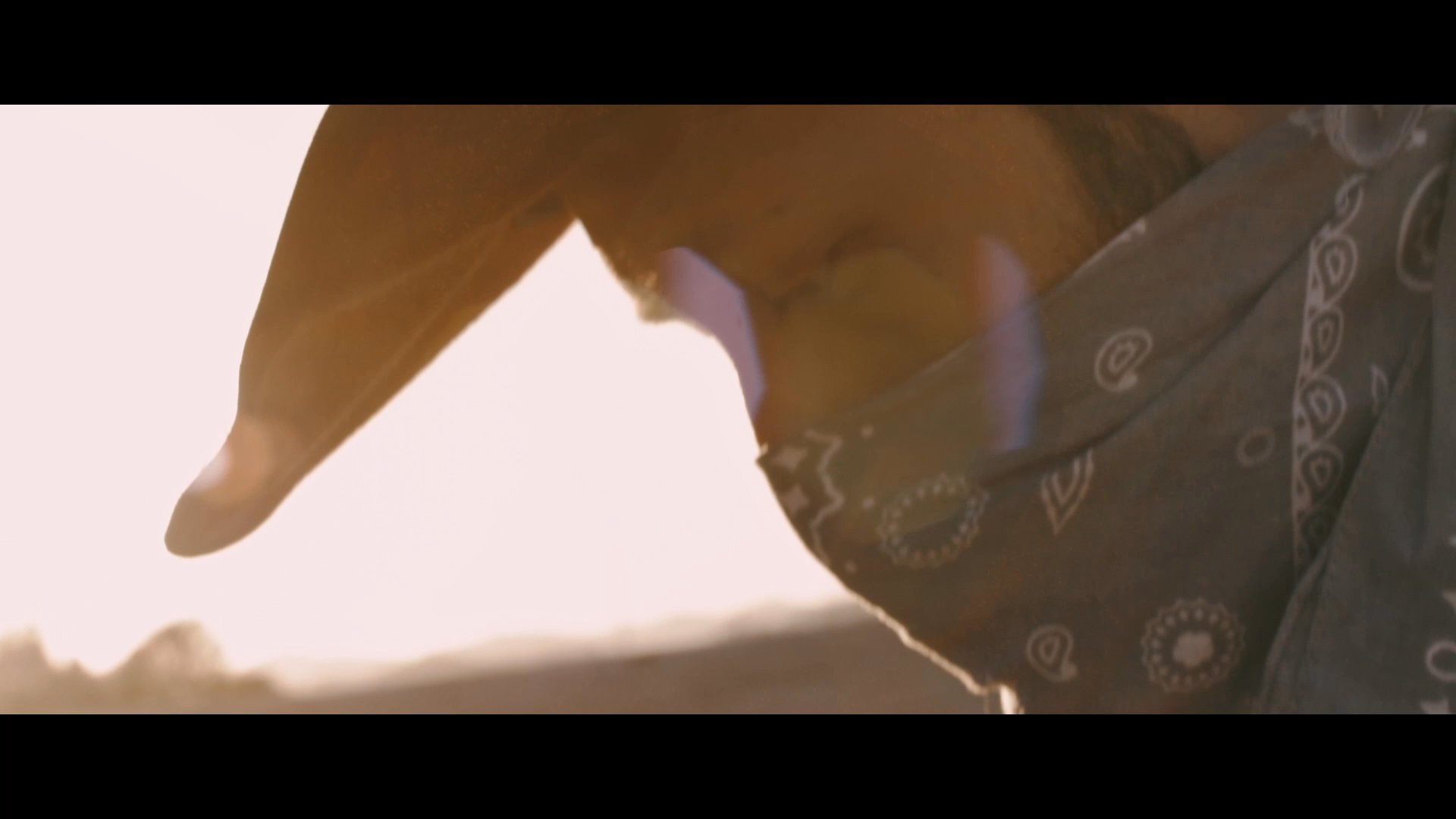 A person pauses in a sun-drenched desert to retrieve a toy sock monkey from a backpack before continuing their journey past a weathered, graffiti-covered wall. The silhouette of the individual moves across the horizon as the sun sets behind them.