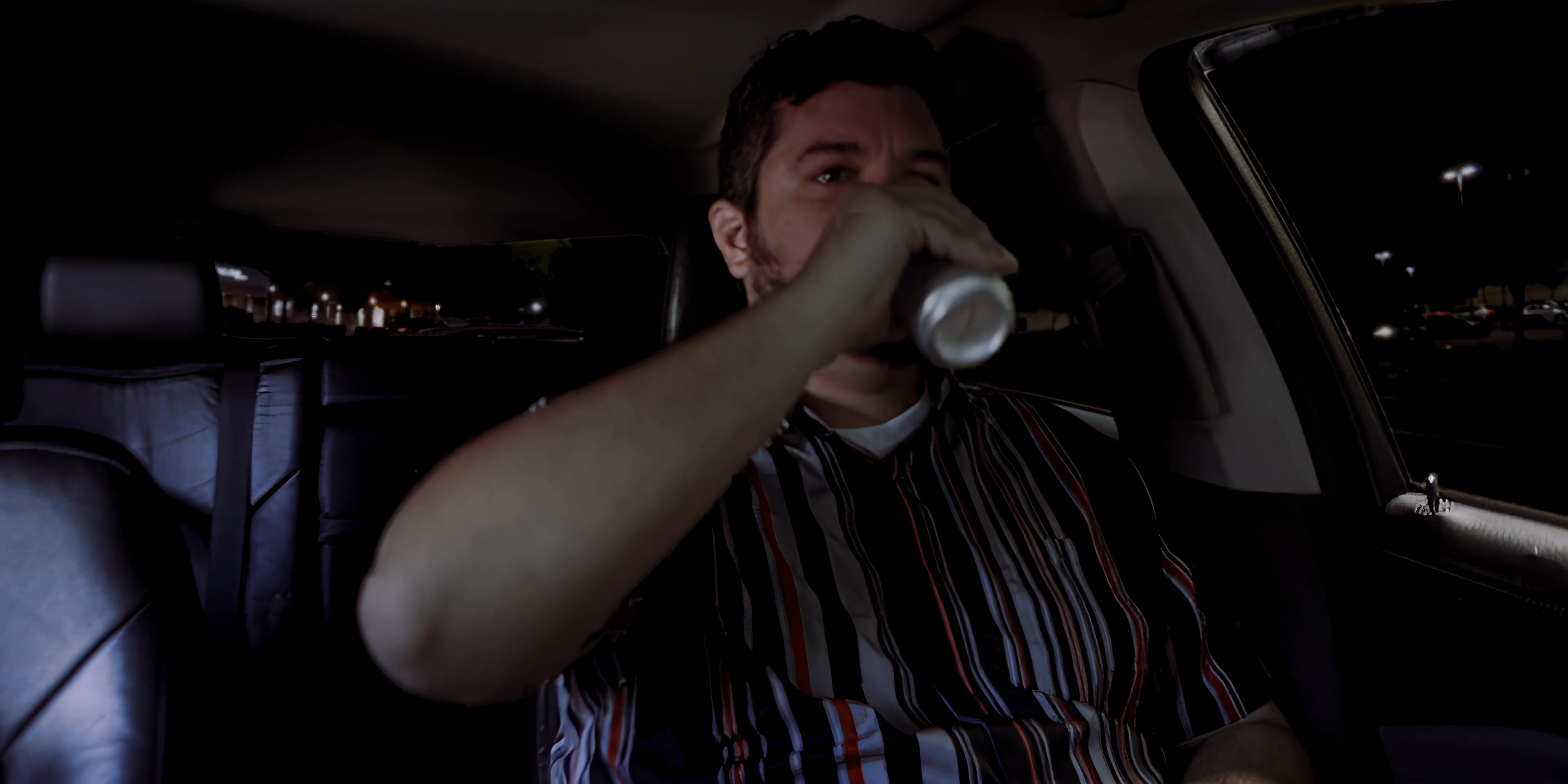 A man sits alone in the driver's seat of a car at night, opening and drinking from a beverage can.
