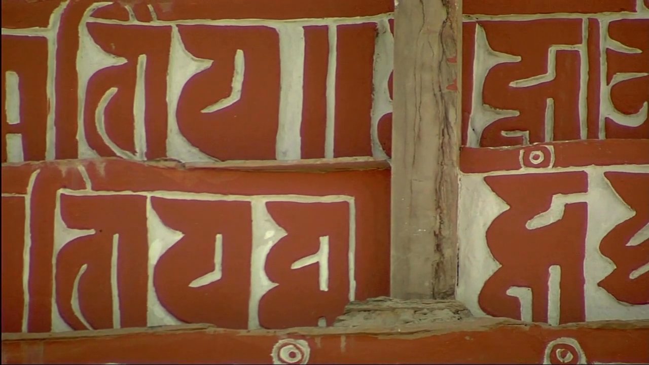 Two individuals engage in a thoughtful conversation while visiting an ornate monastery adorned with traditional carvings and Tibetan script.