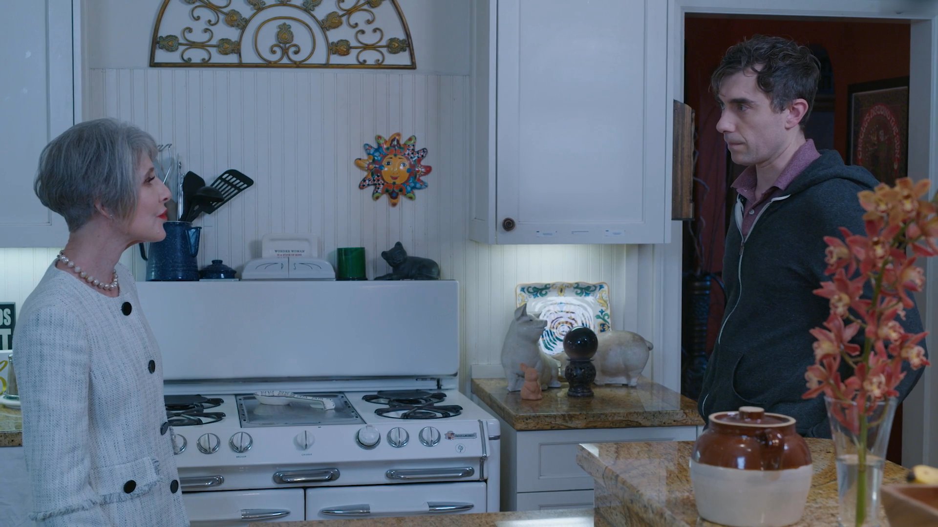 An older woman in a kitchen engages in a serious conversation with a younger man before beginning to pull on yellow rubber cleaning gloves.