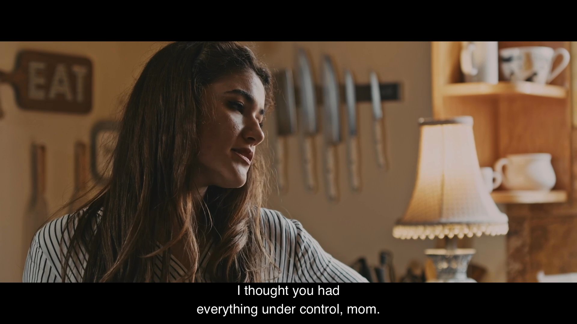 A woman comforts an elderly relative before entering a tense dialogue with her daughter in a kitchen setting. The exchange highlights familial strain and unresolved interpersonal issues.