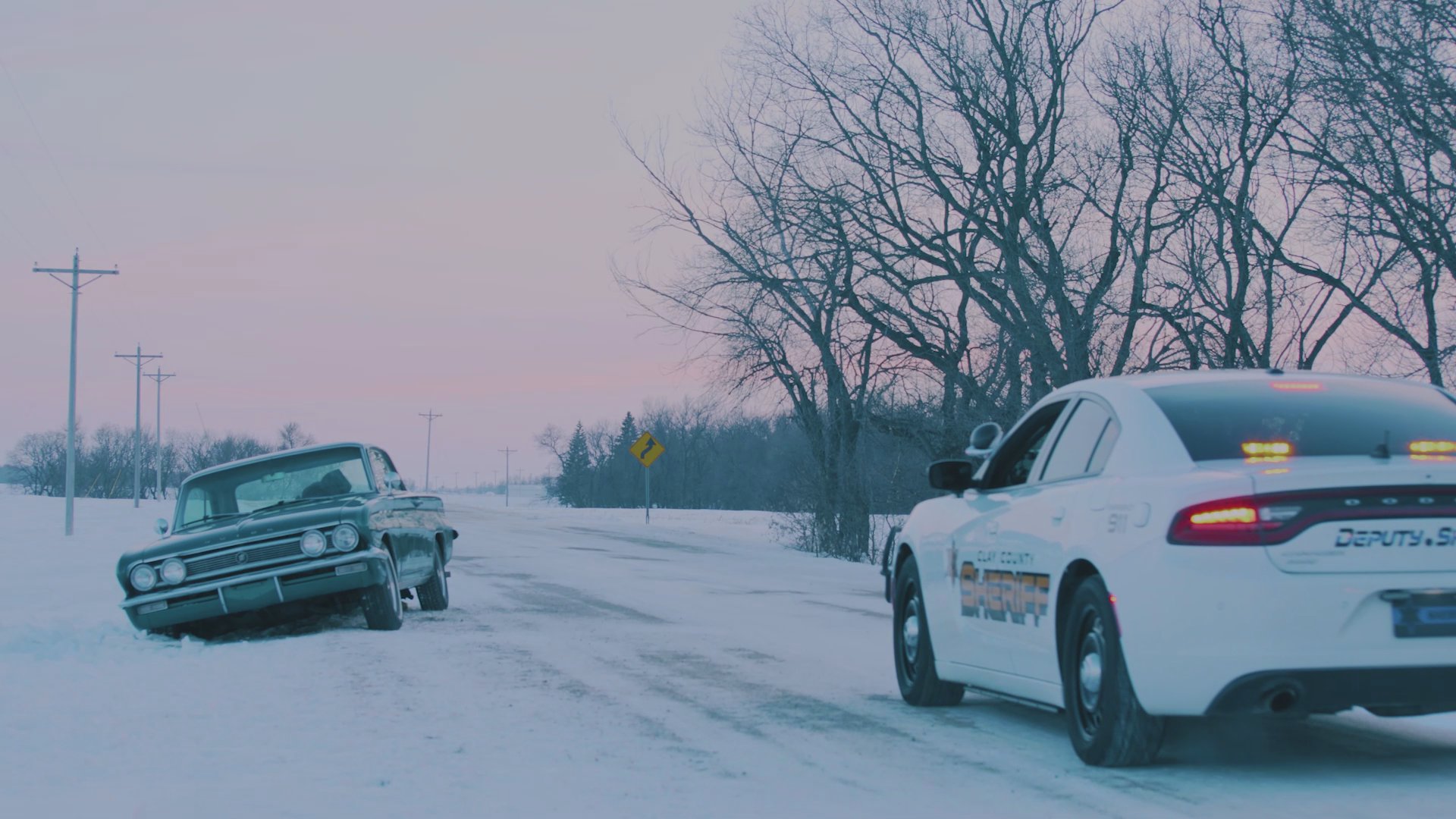A deputy sheriff pulls over a vintage vehicle on a snow-covered rural road, initiating a roadside traffic stop.