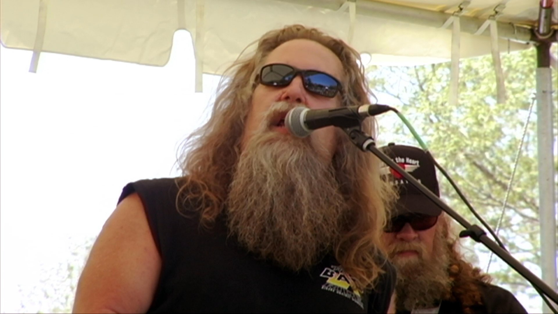 A long-haired, bearded vocalist performs on an outdoor stage, accompanied by a drummer and musical instrumentation.