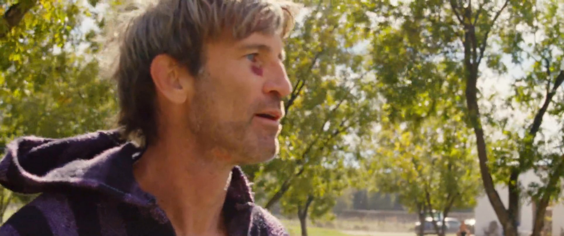 Two men stand outdoors in a tree-lined area, engaging in a serious, focused dialogue. Their facial expressions and body language suggest a tense or significant exchange.