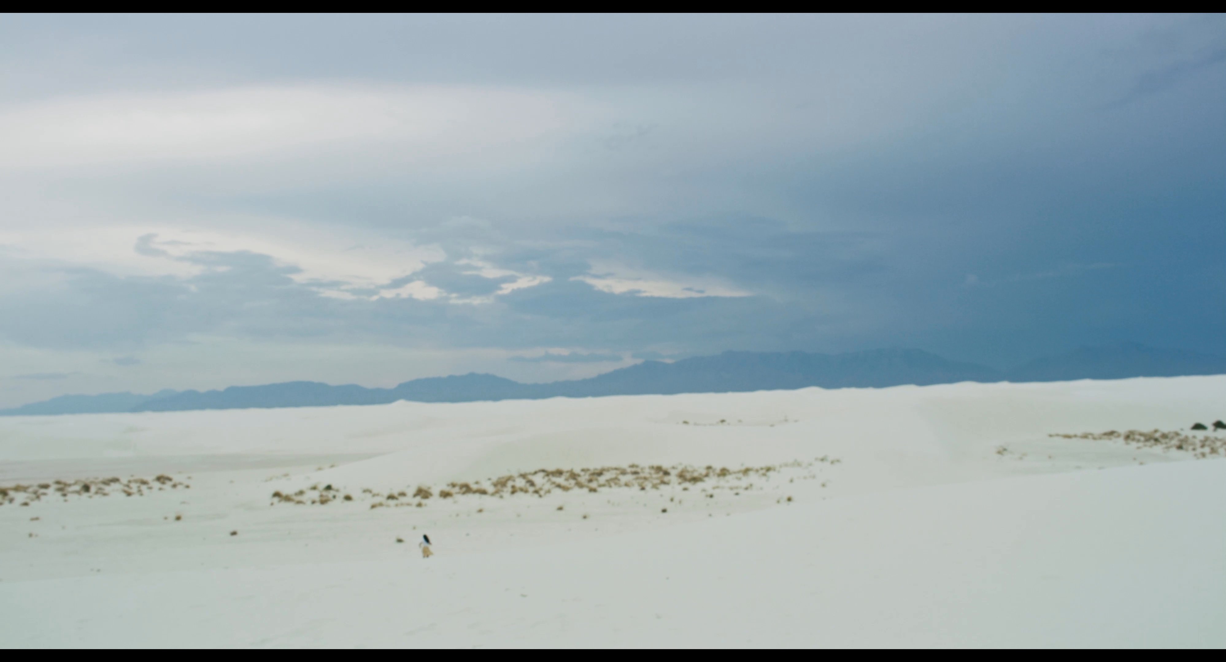 A man and woman navigate a vast white desert landscape before waiting together on a roadside bench with their luggage. The scene highlights a moment of transition during their travel journey.