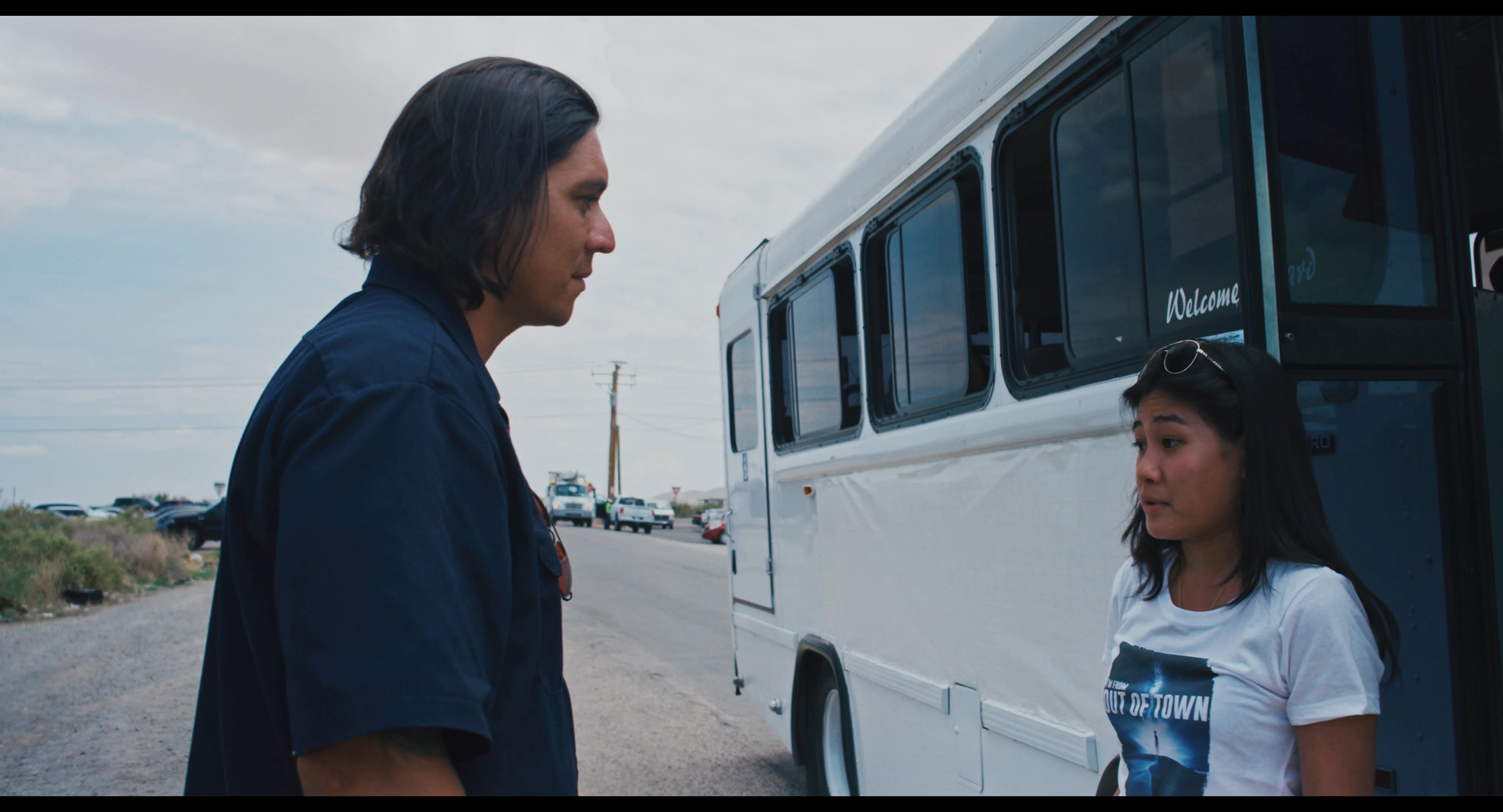 A man and a woman engage in a serious discussion while standing next to a bus on an open, paved lot.