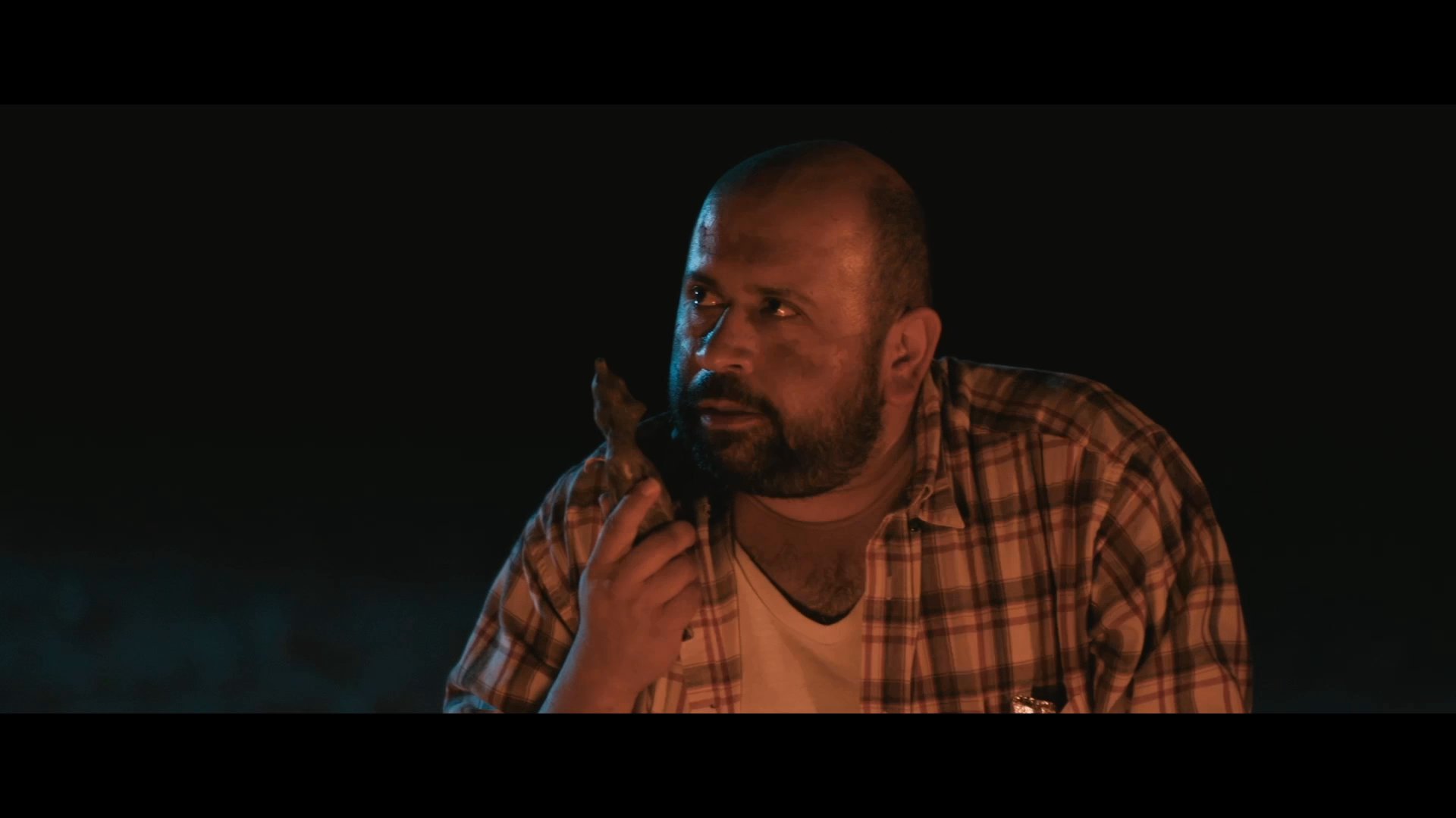 A man sits outdoors at night, entertains others by telling a joke, and uses expressive gestures to deliver the punchline.