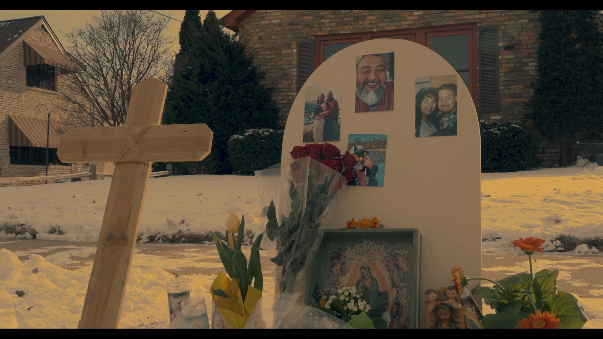 A woman and a young girl visit an outdoor roadside memorial to pay their respects with fresh flowers. They honor a loved one amidst a cold, wintry setting decorated with photos and religious imagery.