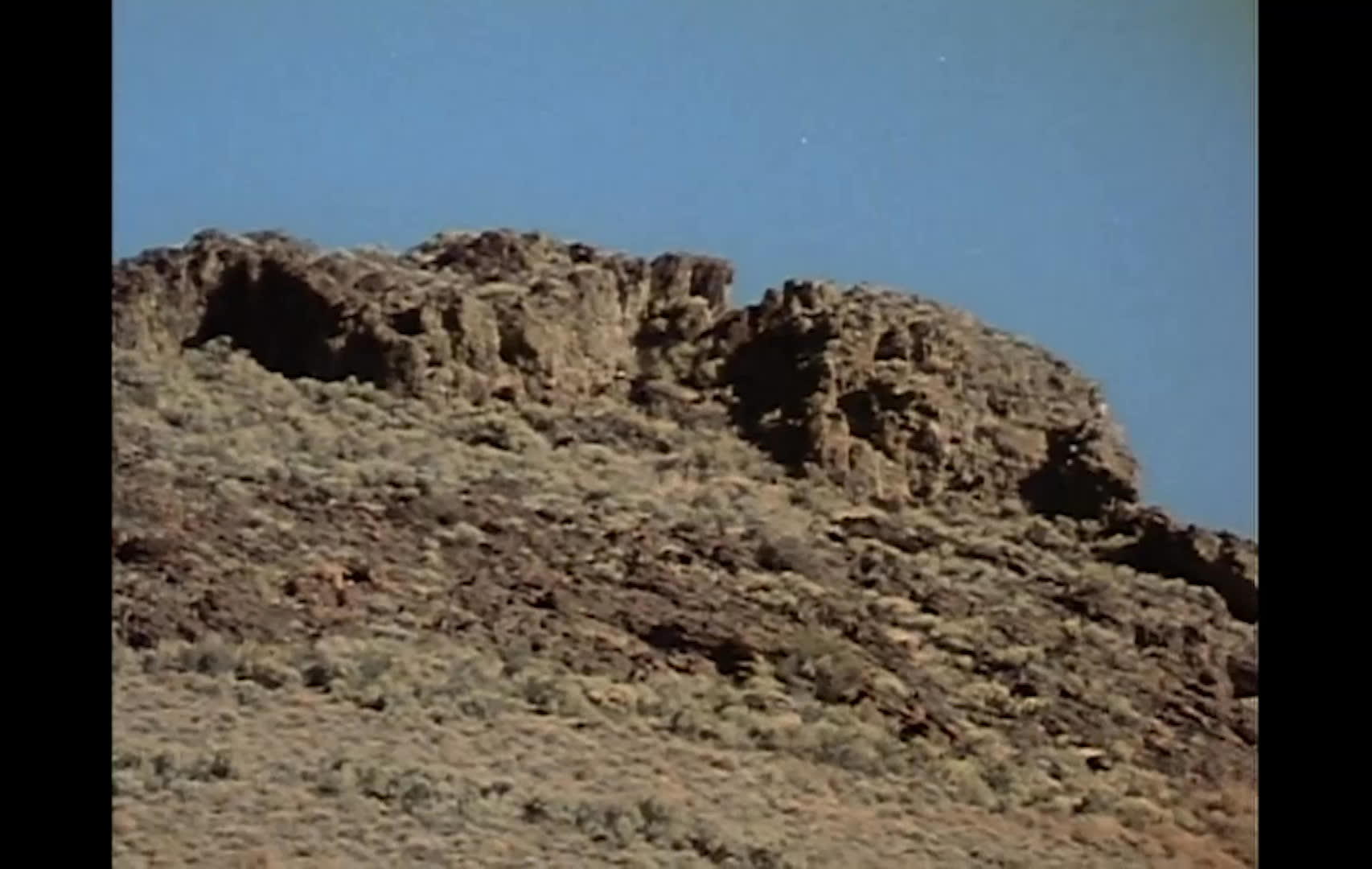 A wide, rugged high-desert landscape surrounds Alkali Cow Camp near Badger Mountain, with a vehicle in the distance and a rustic ranch gate in the foreground. The narration explains that cow camps are places where buckaroos stay, not camps for cattle.