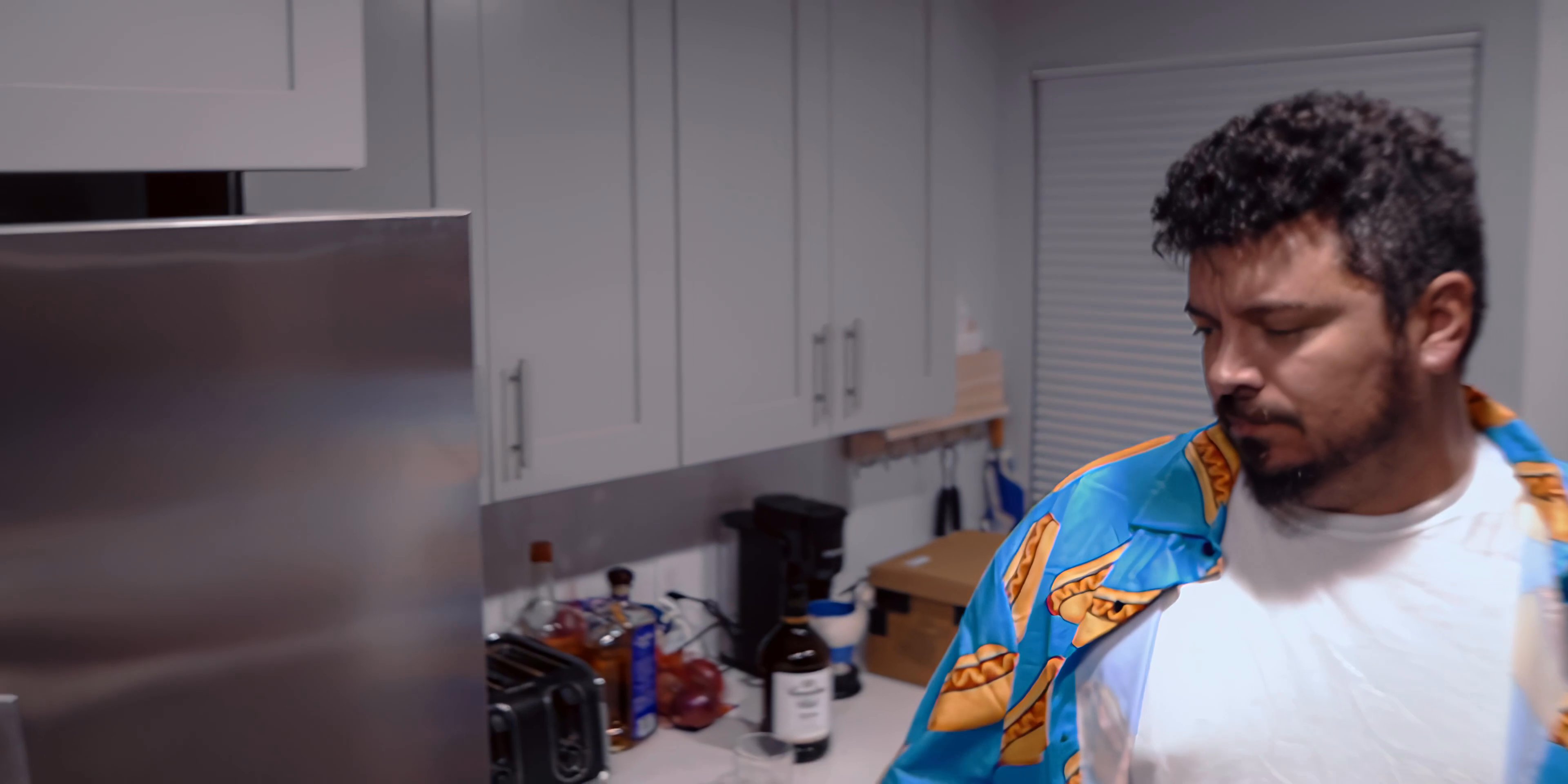 A man in a patterned shirt retrieves a bottle from his kitchen refrigerator and drinks from it. He then proceeds to open the refrigerator to take out a water bottle and consumes it.