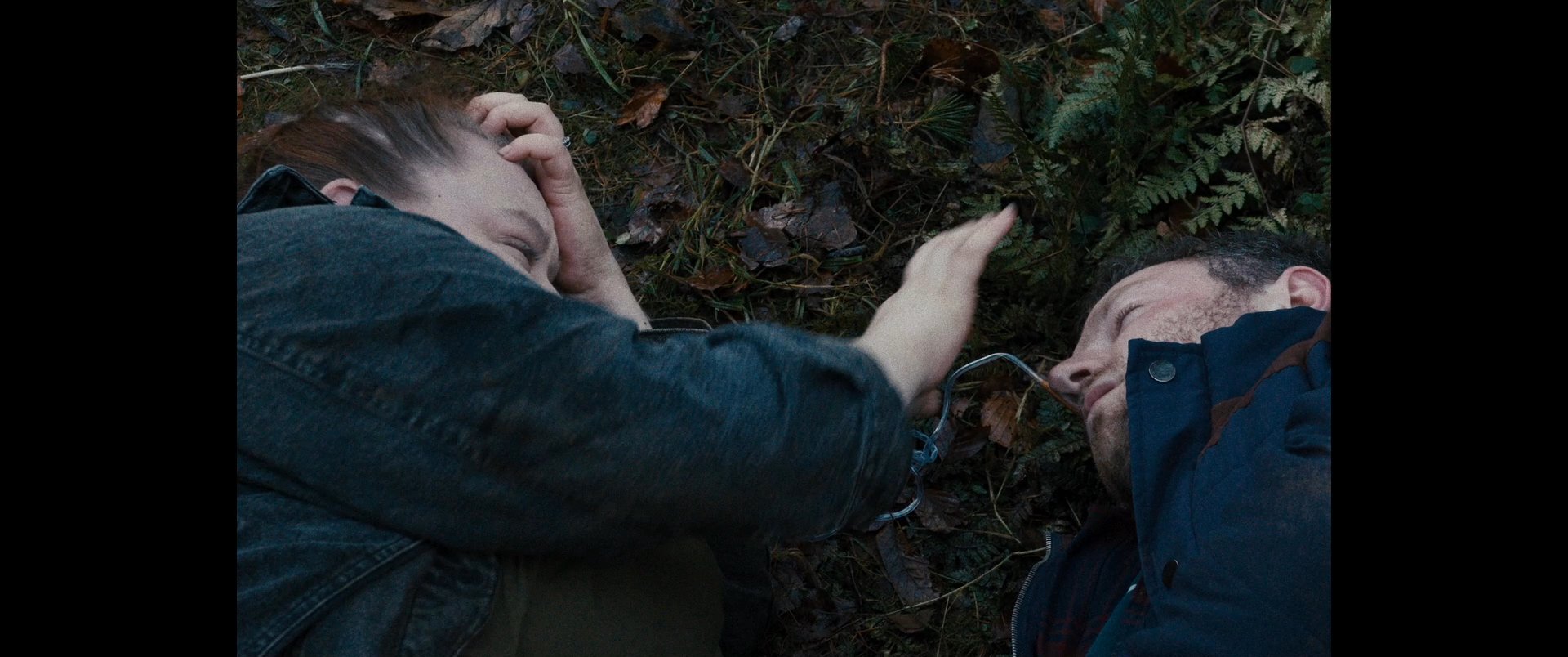 Two individuals share a tender, intimate moment lying together on the forest floor before the scene transitions to a person thoughtfully crafting a textile piece indoors. The contrast highlights moments of interpersonal connection and personal contemplation.