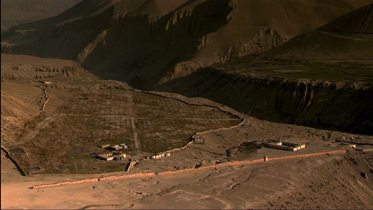 Buddhist monks in traditional red robes walk along stone walls in a remote, mountainous region. The footage transitions to panoramic views of vast, rugged valleys and snow-capped Himalayan peaks.