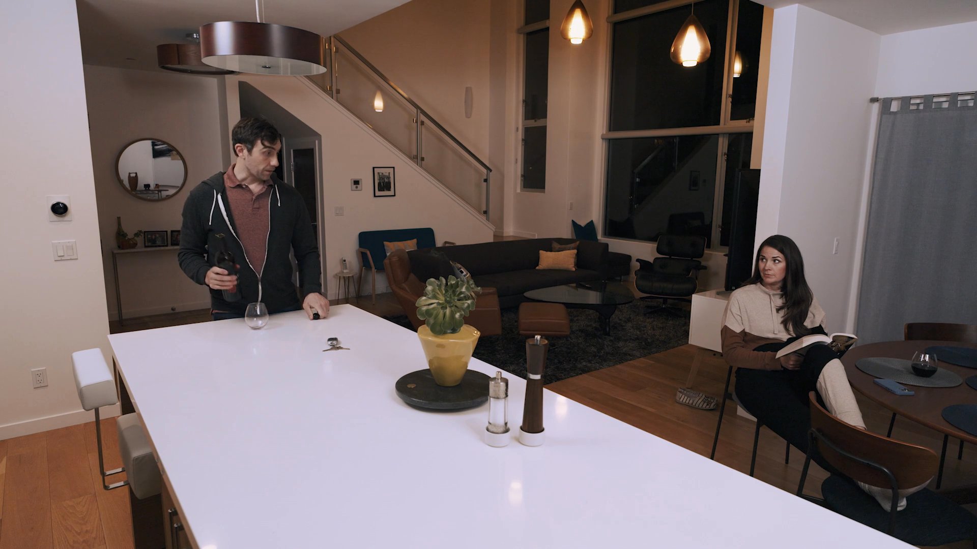 A man moves around a large dining island while a woman sits nearby reading, capturing a quiet, atmospheric moment in a contemporary residence.