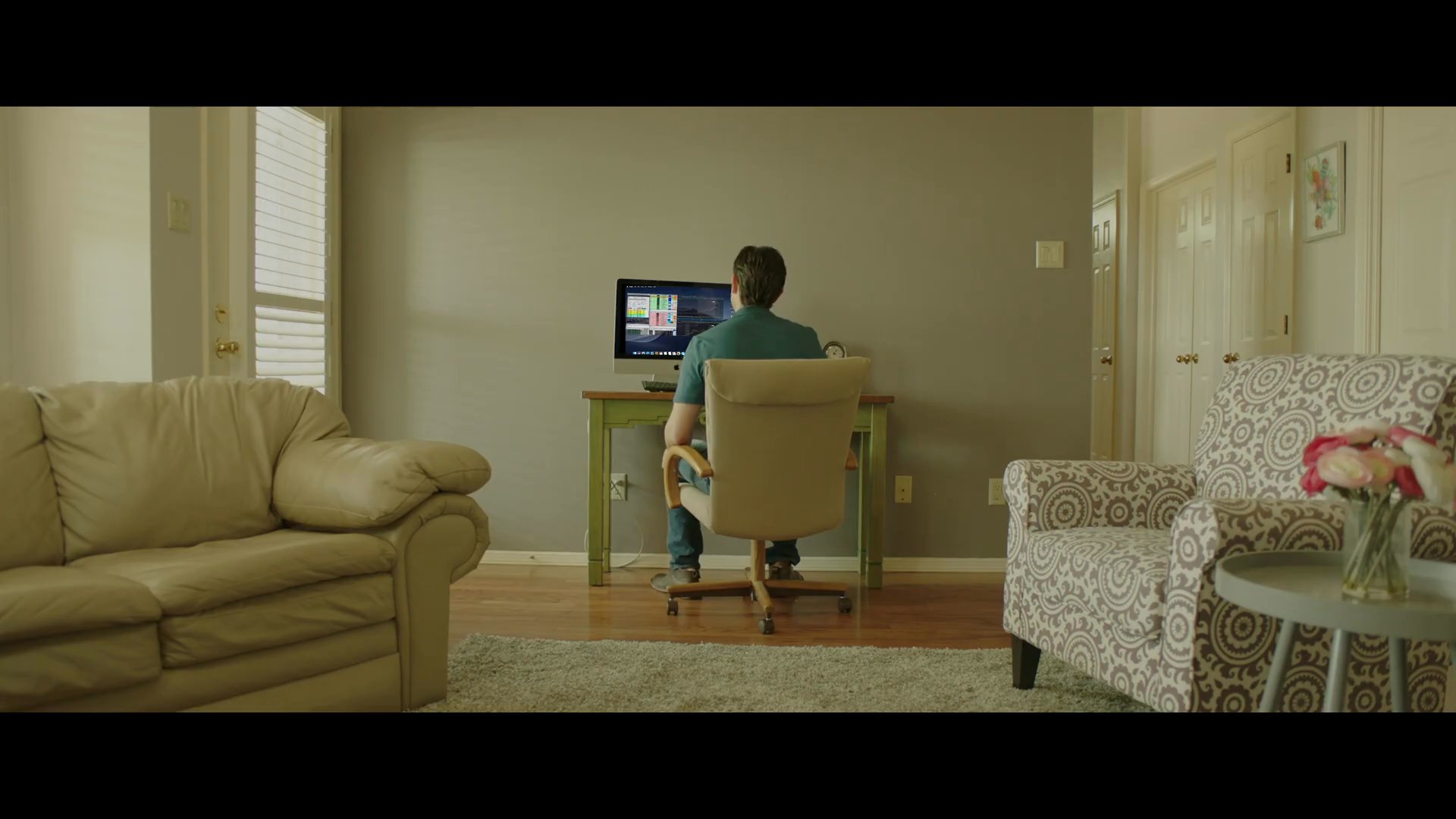 A man monitors complex stock market data on his computer while balancing time with his family at home. He focuses intently on real-time financial charts to make informed trading decisions.