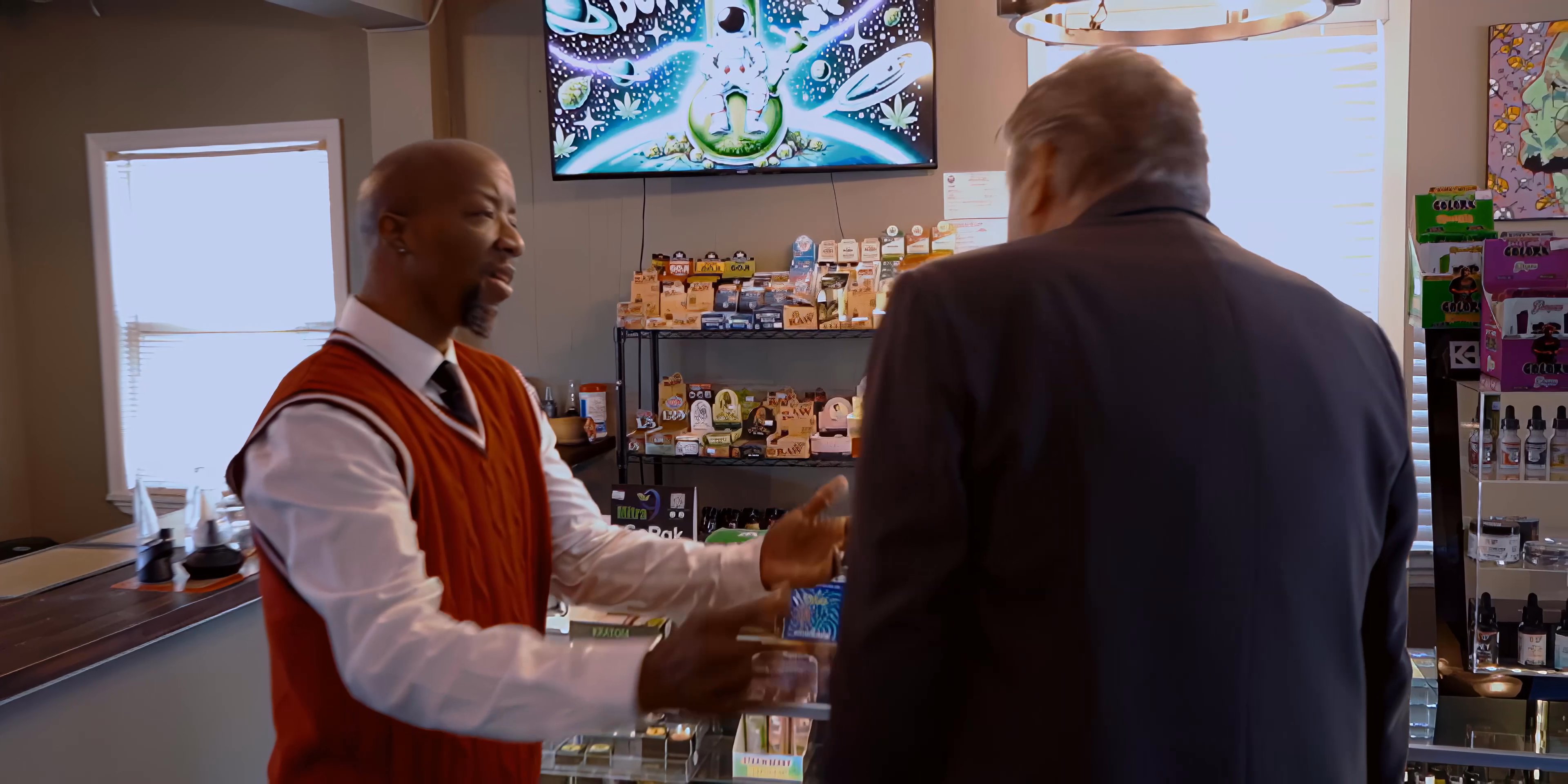 A man in a business suit enters a retail shop and engages in a warm, professional greeting with an employee before transitioning to a workplace discussion.