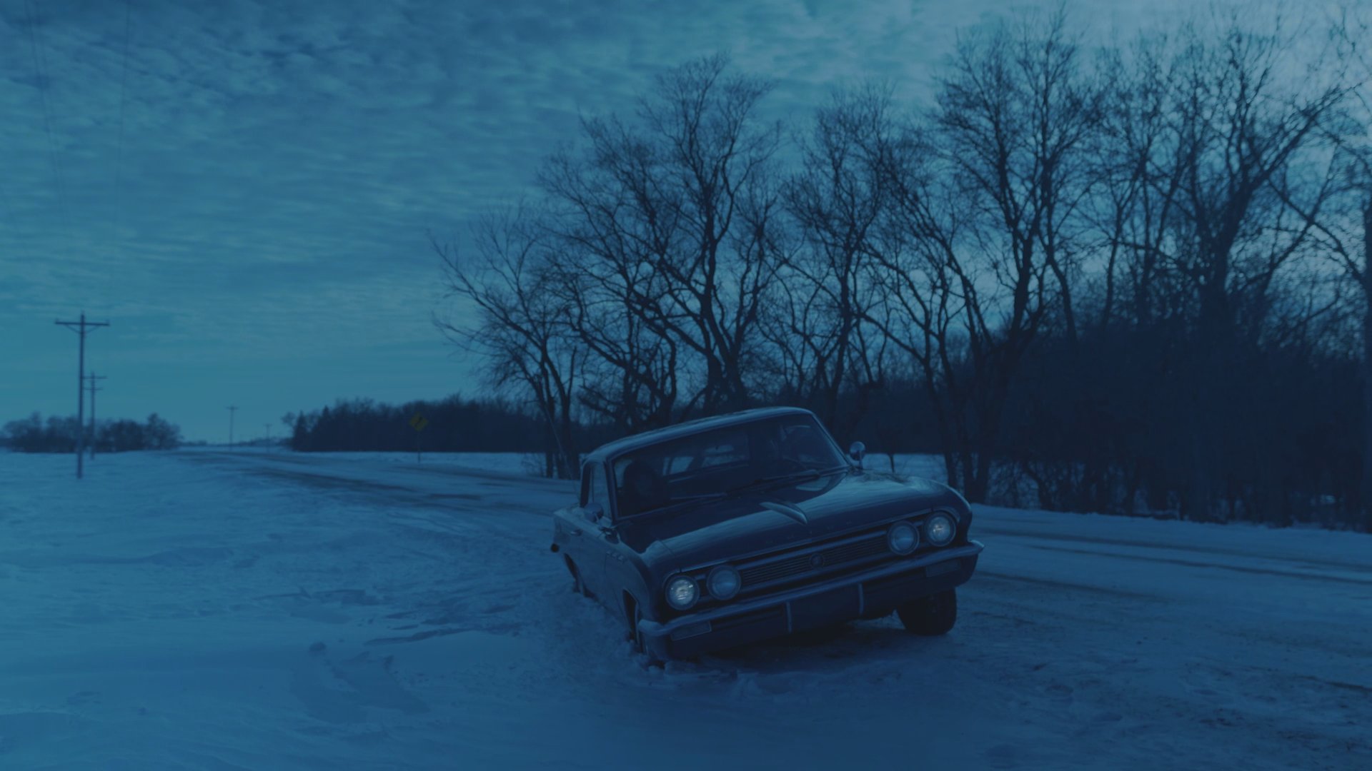 A vehicle remains parked on the side of a snow-covered, desolate road in dim, wintry light while two occupants sit inside. The scene emphasizes an atmosphere of quiet isolation within a frozen landscape.
