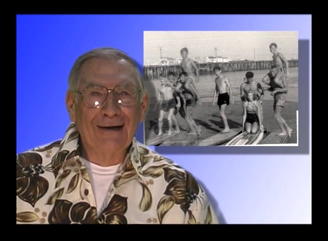 An elderly gentleman shares personal memories of surfing while a vintage photograph of beach activity appears on screen. The interview segment highlights reflections on historical youth experiences.