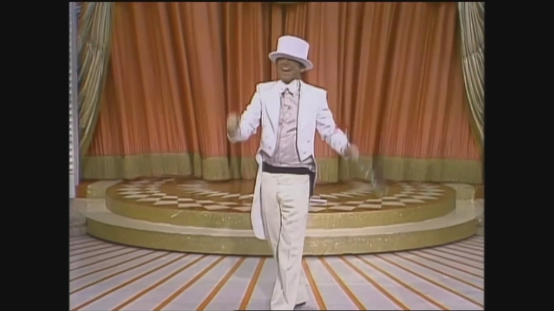 A performer dressed in a white tuxedo and top hat executes a theatrical musical routine on the stage of The Gong Show. The act incorporates classic vaudevillian showmanship and stage presence.