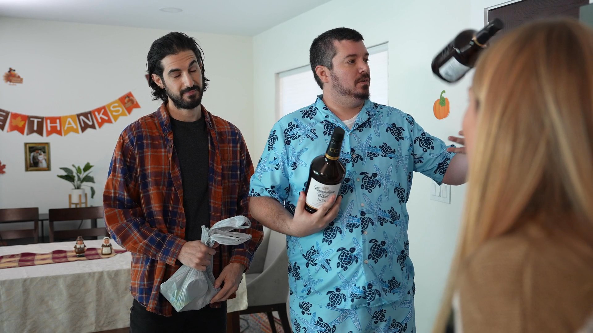 Two men arrive at a house with bottles of wine to join a Thanksgiving gathering, where they are greeted by the hosts in a casual domestic setting.