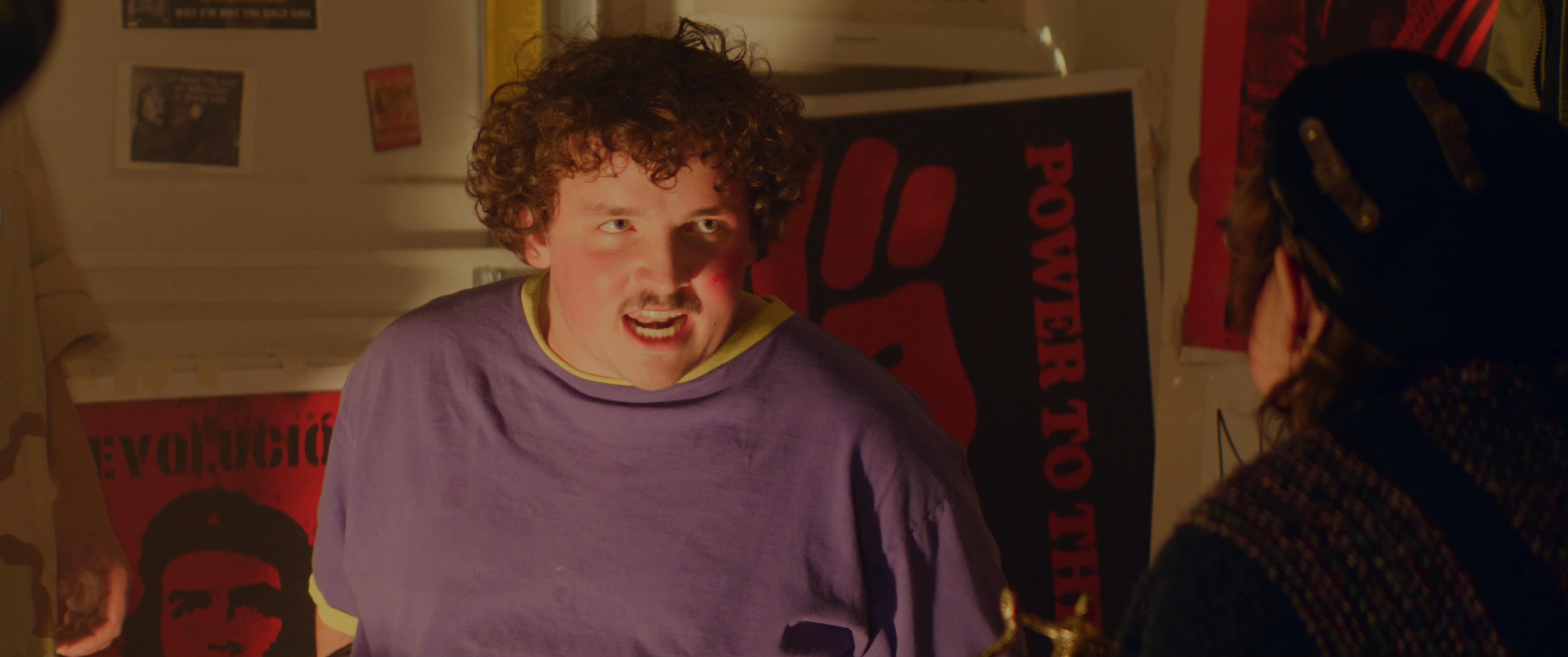 Young men engage in a serious conversation within an room adorned with political posters and imagery. The subjects exchange views while standing in a dimly lit environment characterized by a focused, reflective atmosphere.