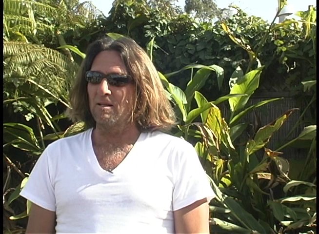 A man wearing a white v-neck shirt and sunglasses stands in a lush outdoor setting, delivering a message to the camera.