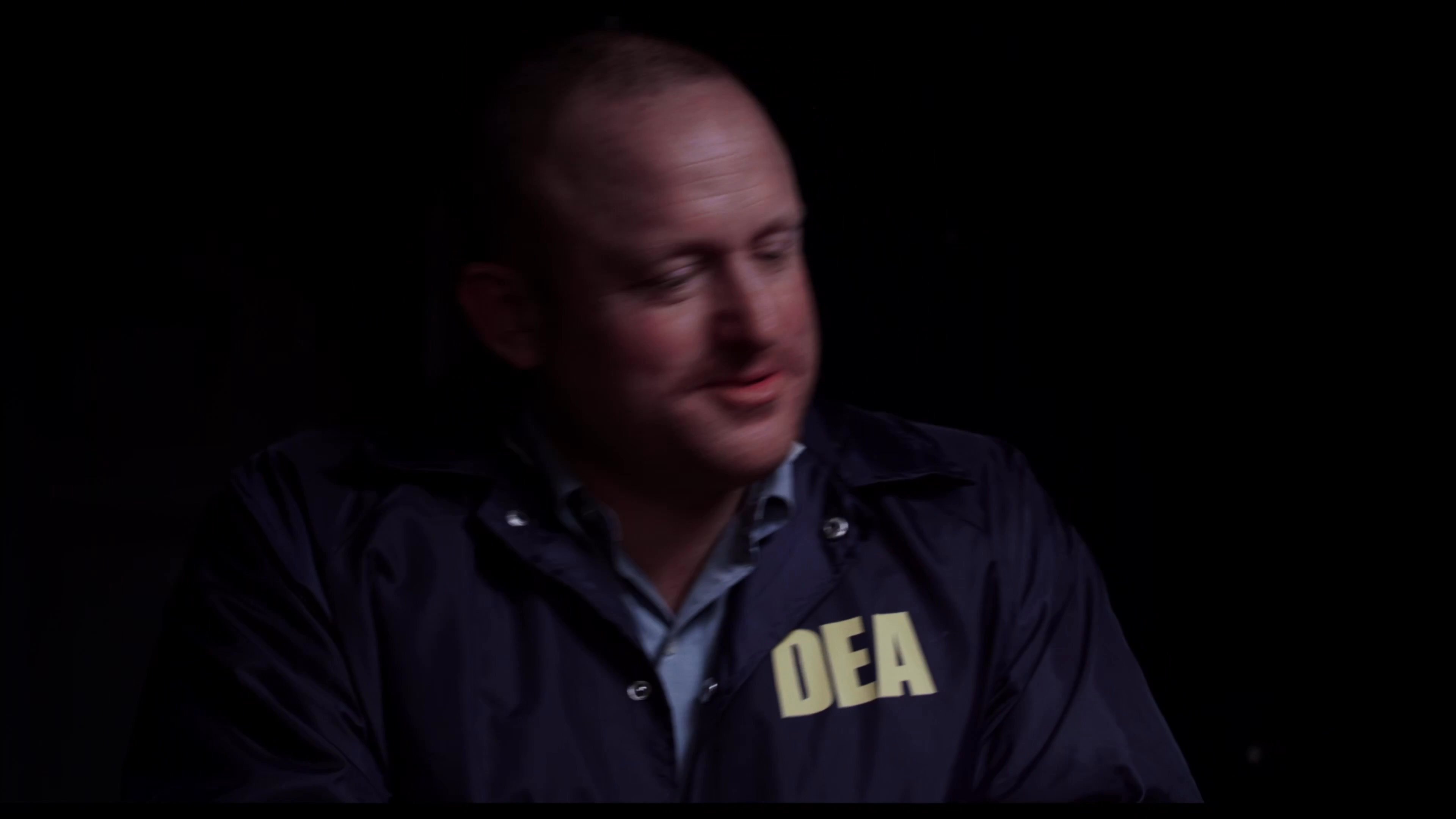 A federal agent conducts an interrogation of two suspects wearing orange detention jumpsuits in a dark, confined setting. The agent maintains a calm demeanor while questioning the handcuffed individuals.