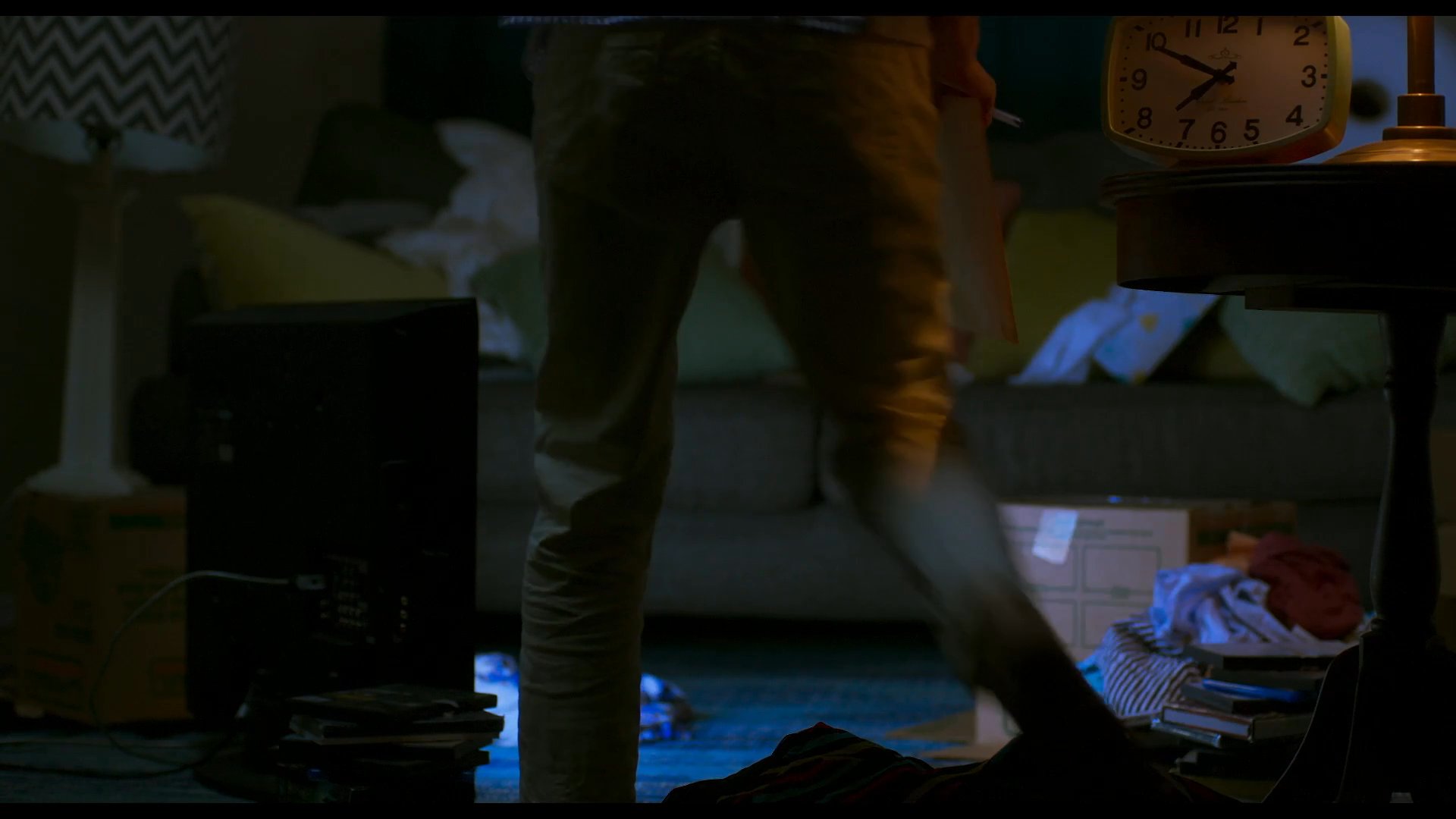 A woman relaxes in a dimly lit living room, transitioning from a kneeling position to sitting on the floor while reviewing a document and drinking from a glass.