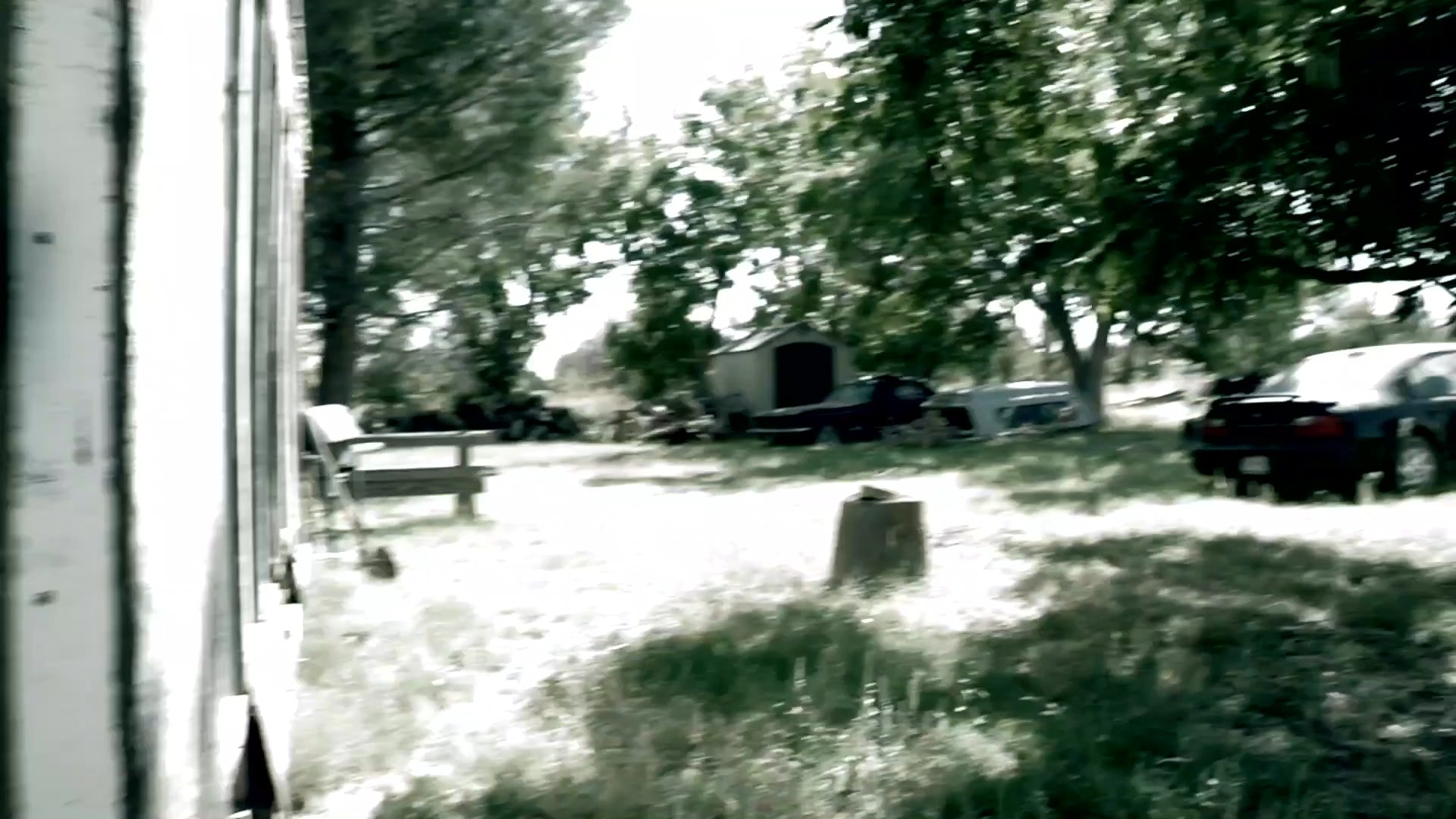 A handheld camera moves through a sunny, grass-covered backyard area adjacent to a residential building. The perspective captures mature trees, parked vehicles, and a rustic outdoor environment during daylight hours.