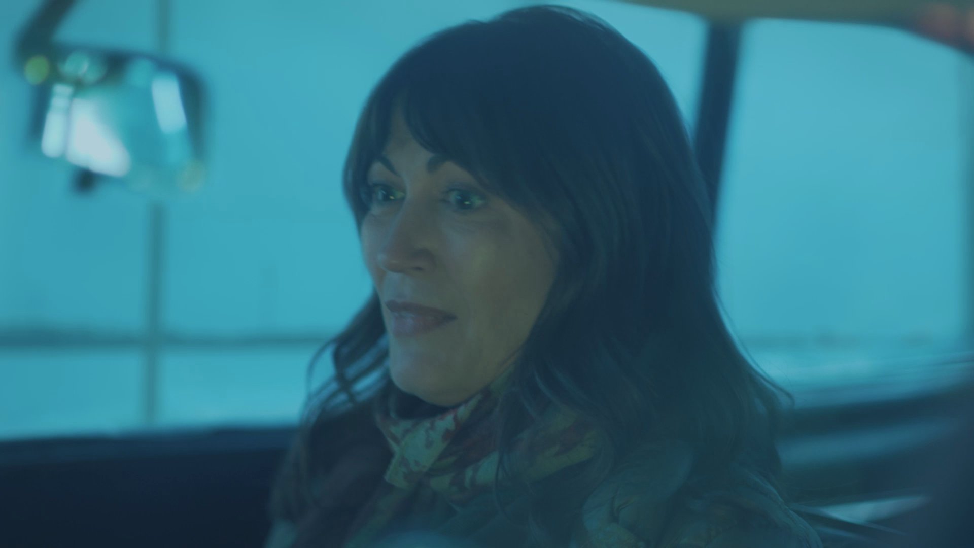 A man and woman engage in a quiet conversation while driving through a dimly lit, atmospheric landscape at night.