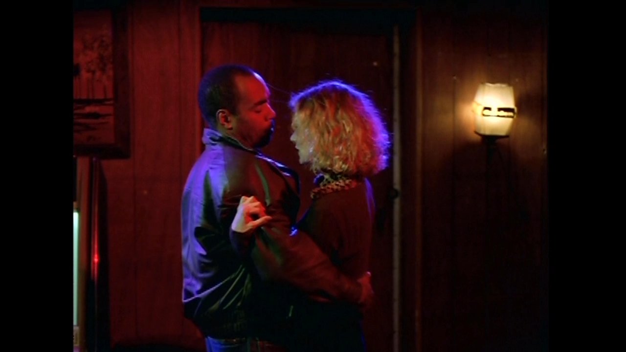 A man and a woman share an intimate, slow dance in a dimly lit, wood-paneled room. The two maintain close physical contact while swaying gently together.