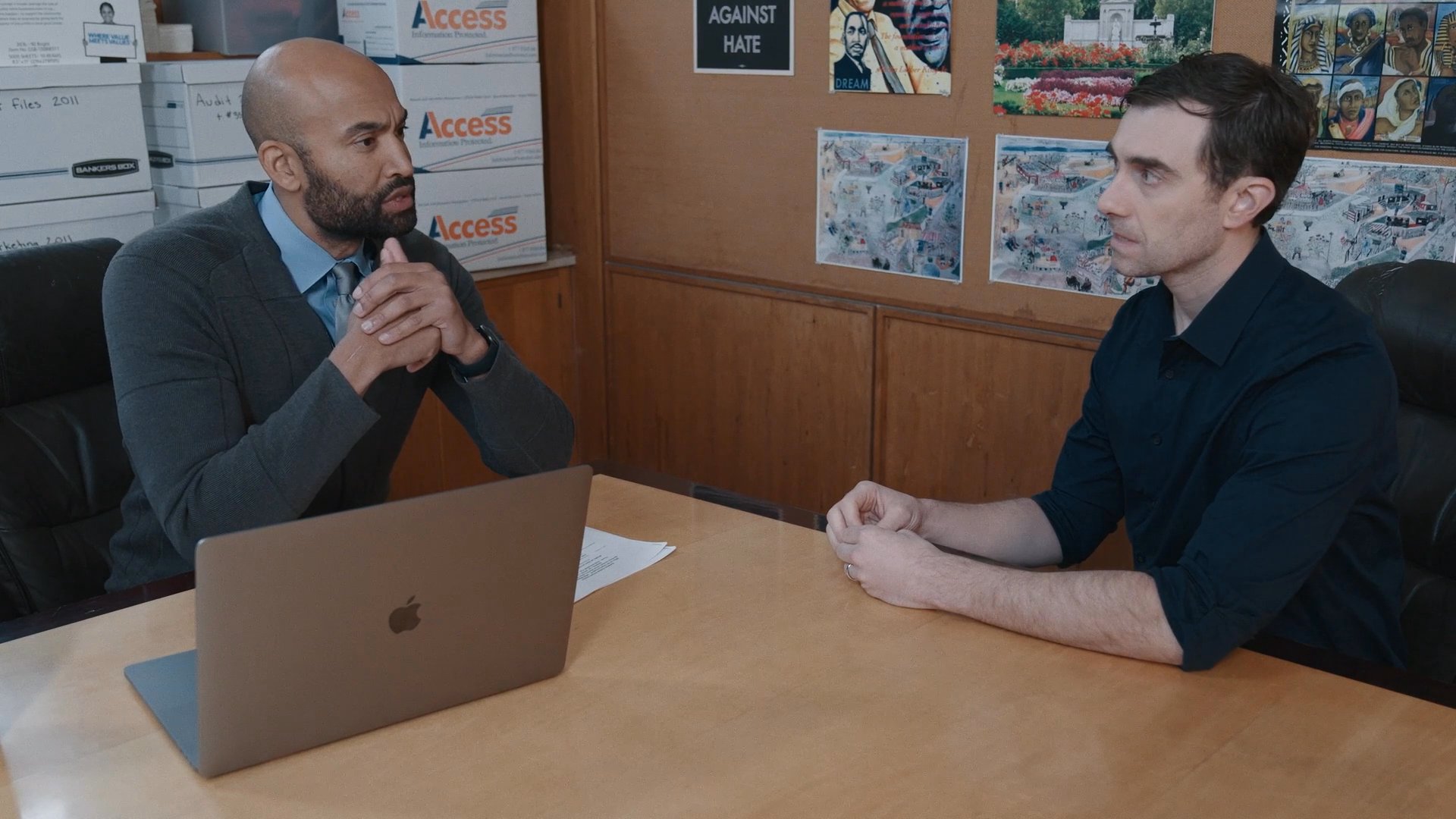 Two men engage in a serious professional discussion while seated at an office desk. They exchange information and perspectives throughout their interaction in a collaborative work environment.