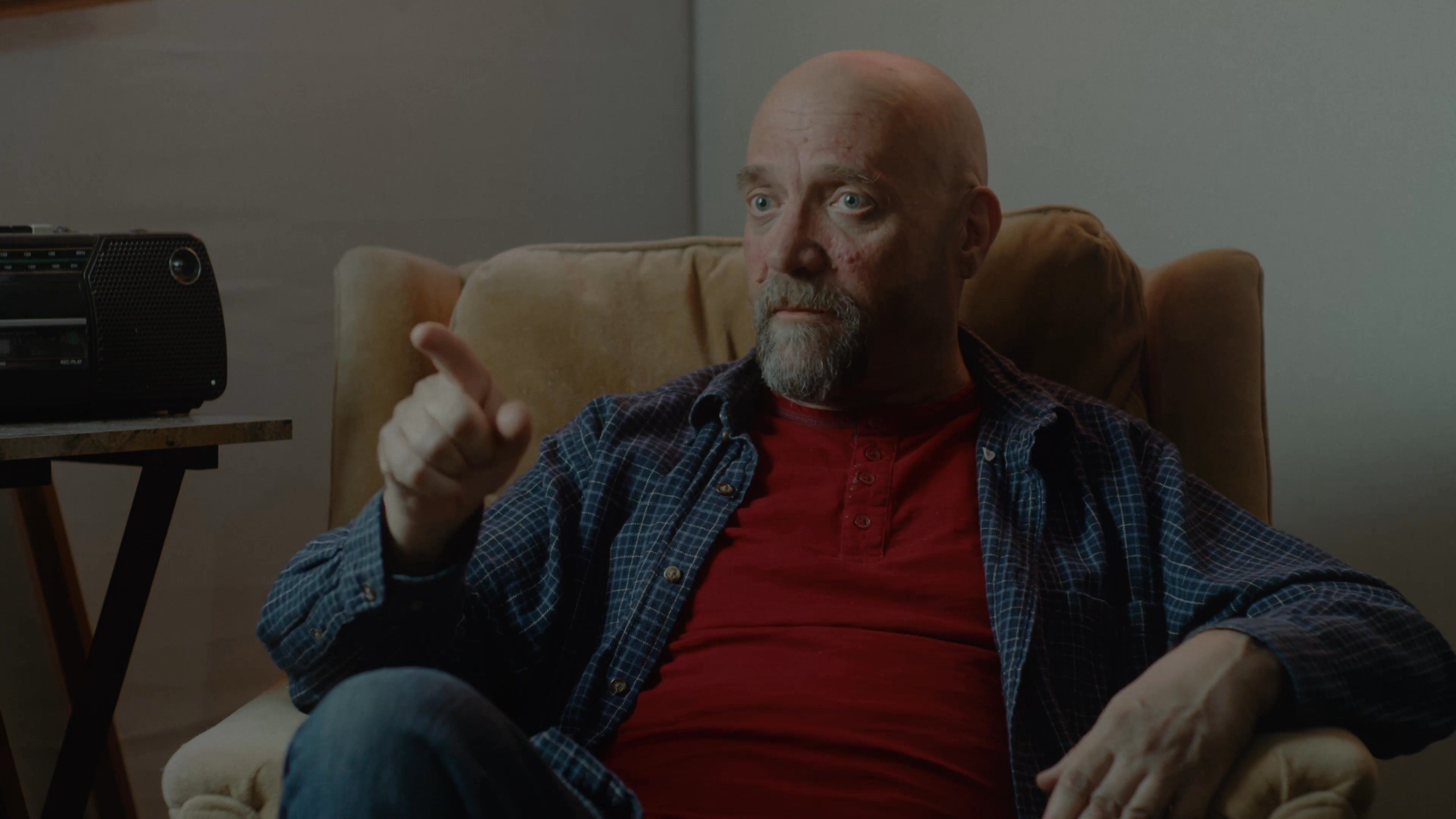 Two men engage in a calm, seated conversation within a casual home living room setting. The participants exchange perspectives while maintaining a relaxed and reflective tone.