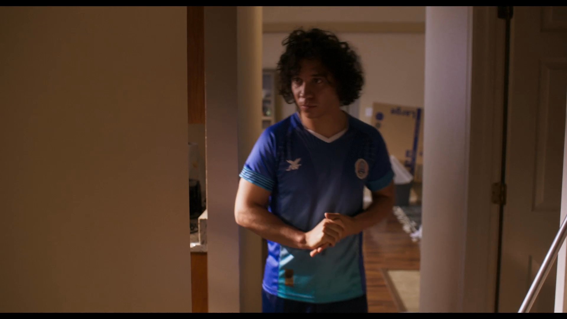 A young man wearing a soccer jersey walks through a residential home toward an adult woman standing in a doorway to engage in a conversation.