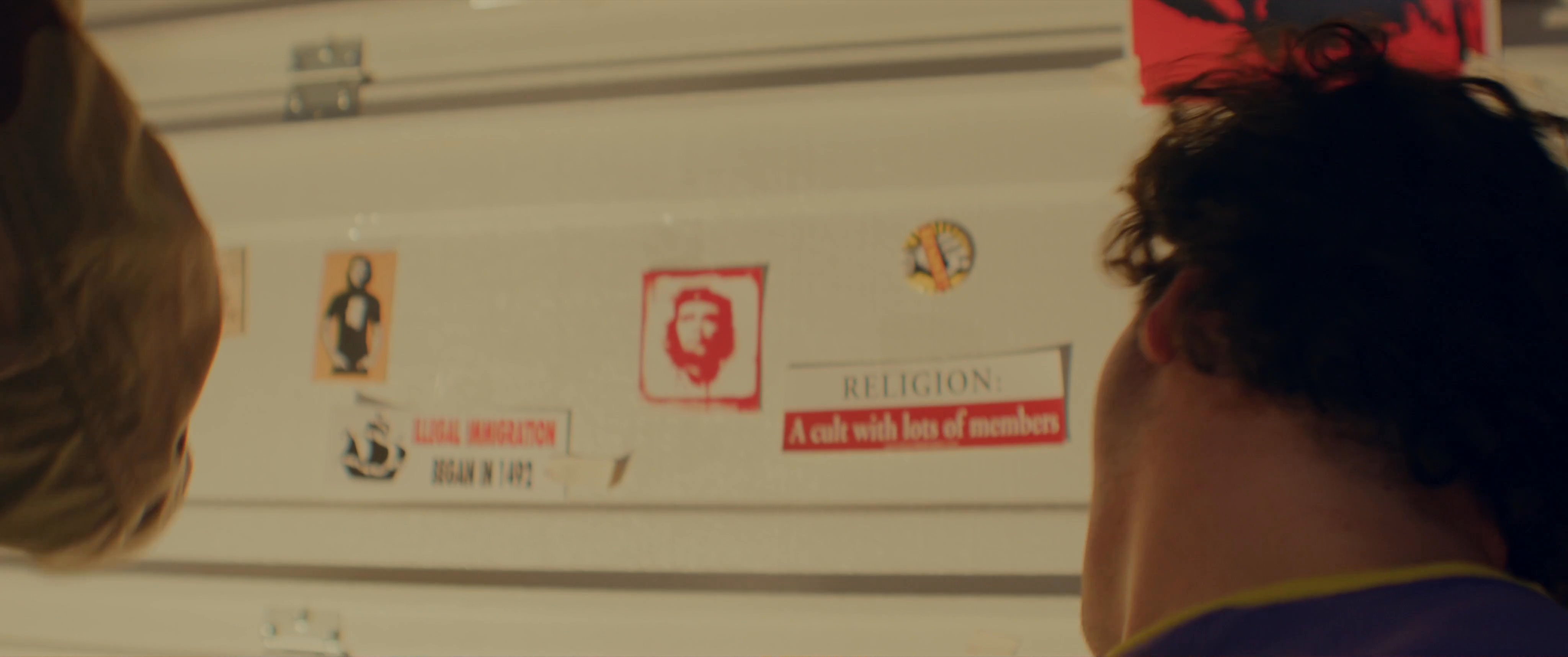 A man wearing red-tinted glasses questions a youth in a room decorated with radical posters. The interaction escalates as a bearded individual enters and introduces a firearm into the tense setting.