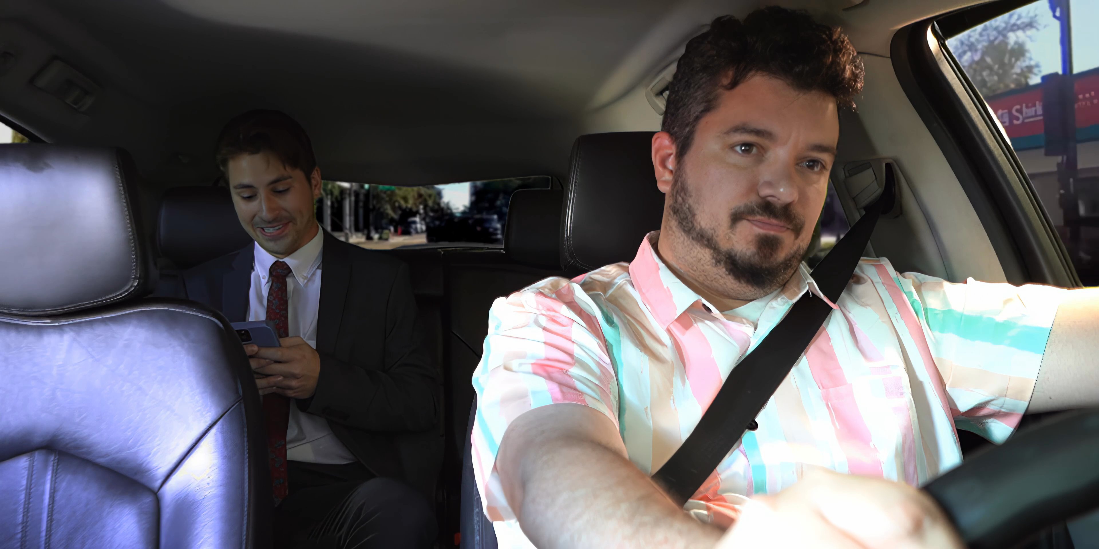 A driver in a patterned shirt navigates a vehicle while engaging in a casual conversation with a passenger sitting in the back seat. Both parties interact periodically while the driver focuses on the road ahead.
