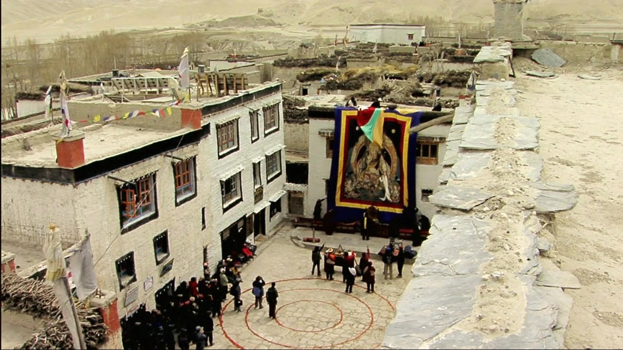 Devotees gather in the courtyard of a Tibetan monastery for a traditional religious ceremony featuring large thangka tapestries and communal ritual activities.