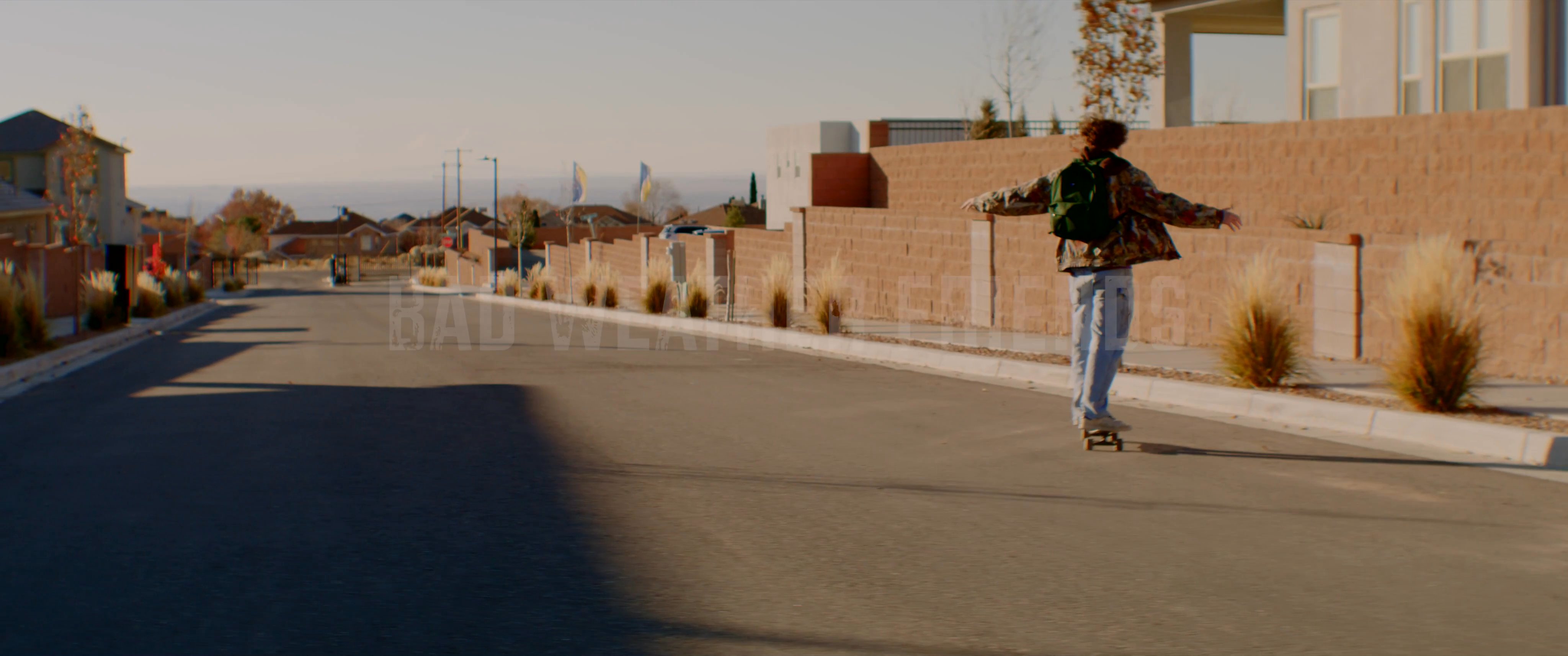 A young individual skateboards through a quiet residential neighborhood while wearing a backpack. The scene transitions to a title card for Bad Weather Friends against a backdrop of distant mountains.