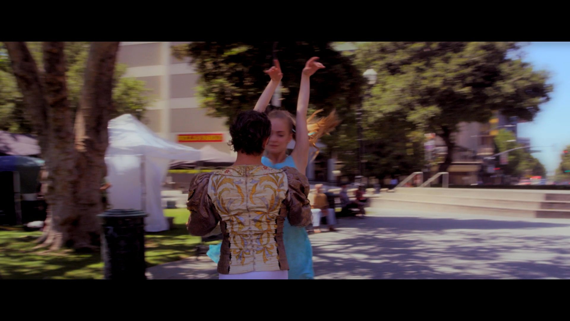 A ballet duo performs an elegant routine in a public park setting, executing synchronized movements and lifts. The sequence highlights their technical precision and artistic expression against an urban backdrop.