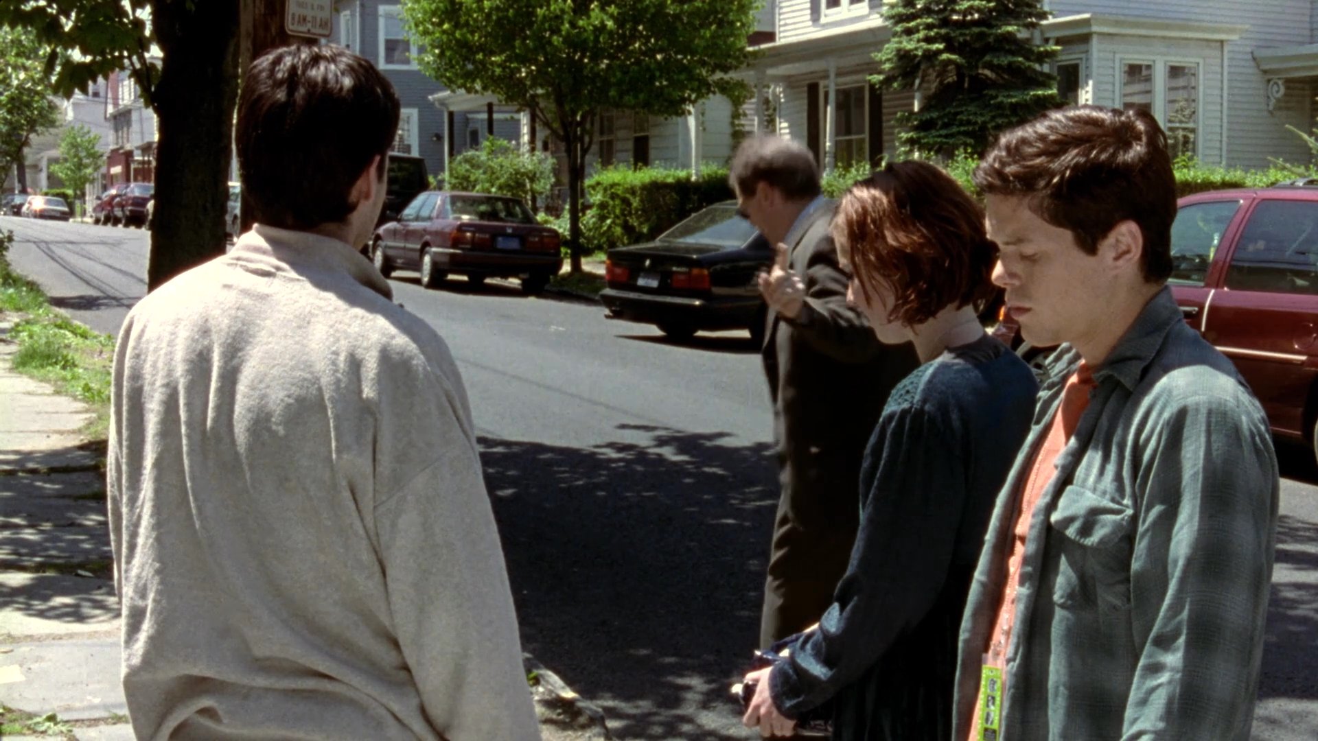 A man in a suit delivers a small, dark package to a group of individuals standing on a suburban residential street. After handing over the item, he walks away while the group remains on the sidewalk.