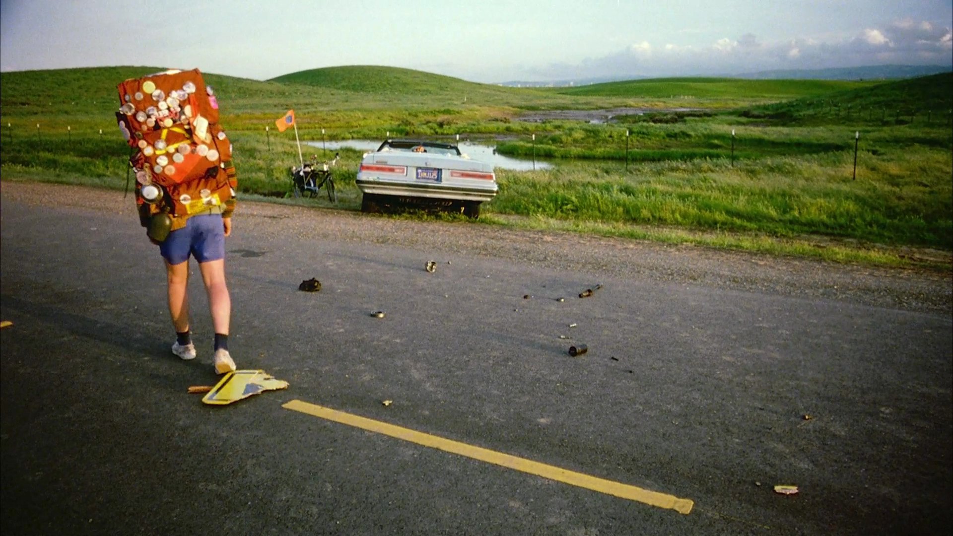 A man wearing a heavily accessorized backpack walks away from a convertible on a rural highway. He appears surprised or disappointed as he looks back toward the vehicle.