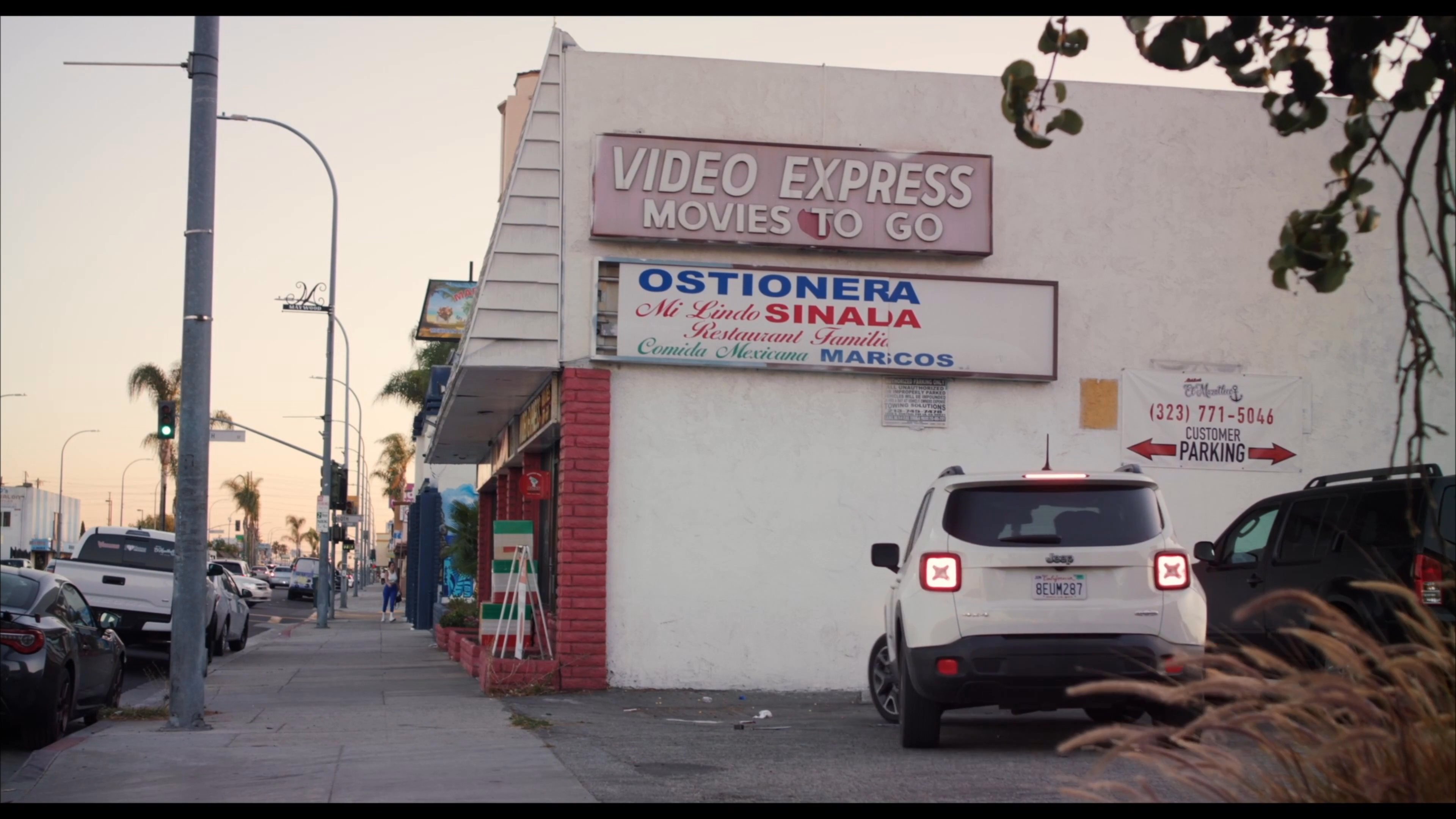 Two men arrive at a neighborhood video rental store after driving through a desert landscape. The scene transitions to the interior where a customer browses shelves of movies under the store lighting.