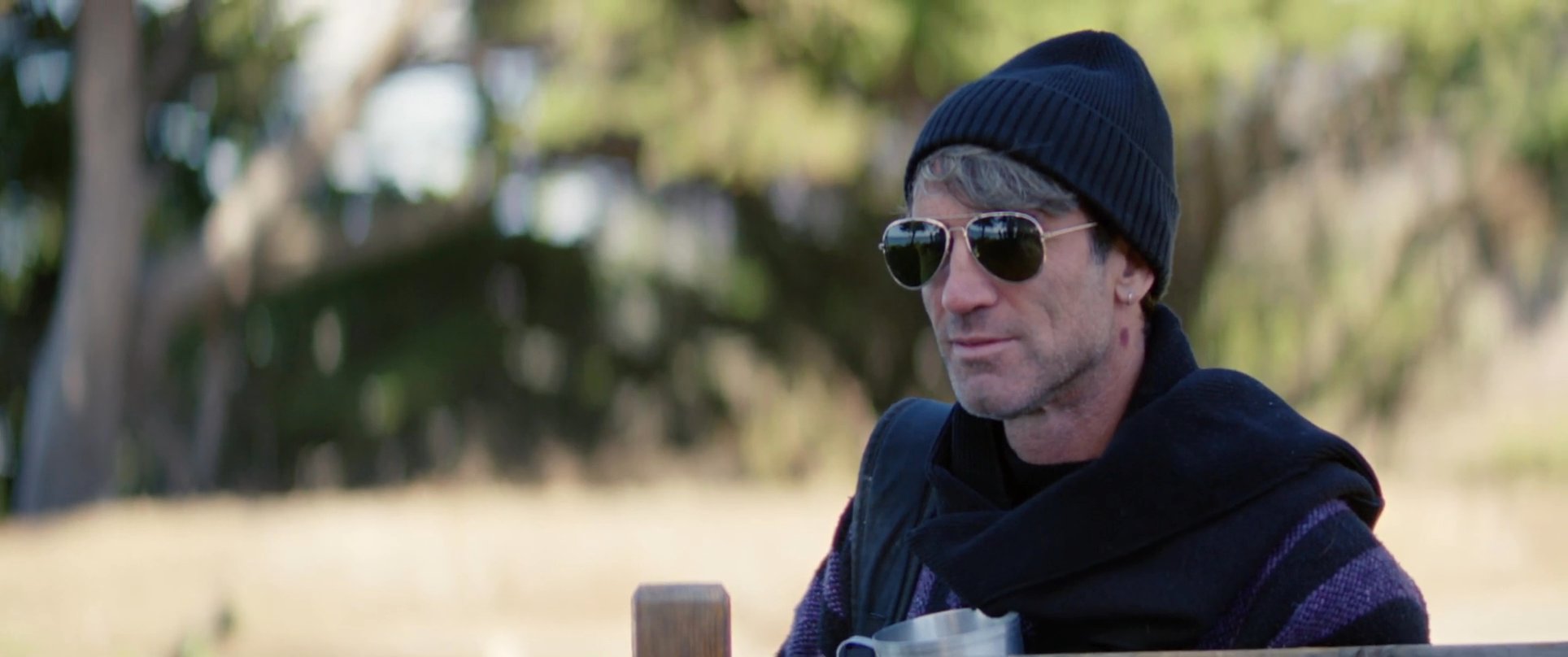 Two men wearing beanies and sunglasses engage in conversation while sitting on a wooden bench overlooking a coastal landscape.